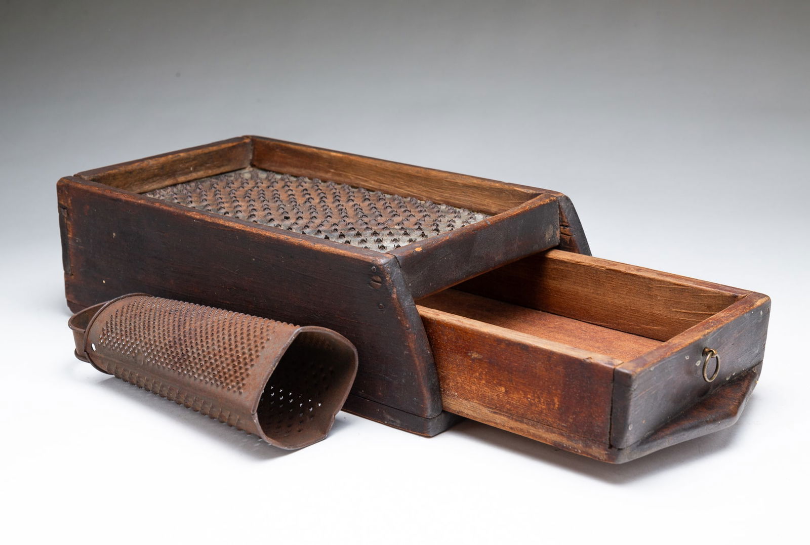 TWO AMERICAN GRATERS.: Mid-19th Century. A wood cased hanging grater, nail and screw construction with a single drawer 15"h. 7"w. 3.75"d Together with a cylindrical grater with handle, 7.5"h.Provenance: From the Lifetime Co