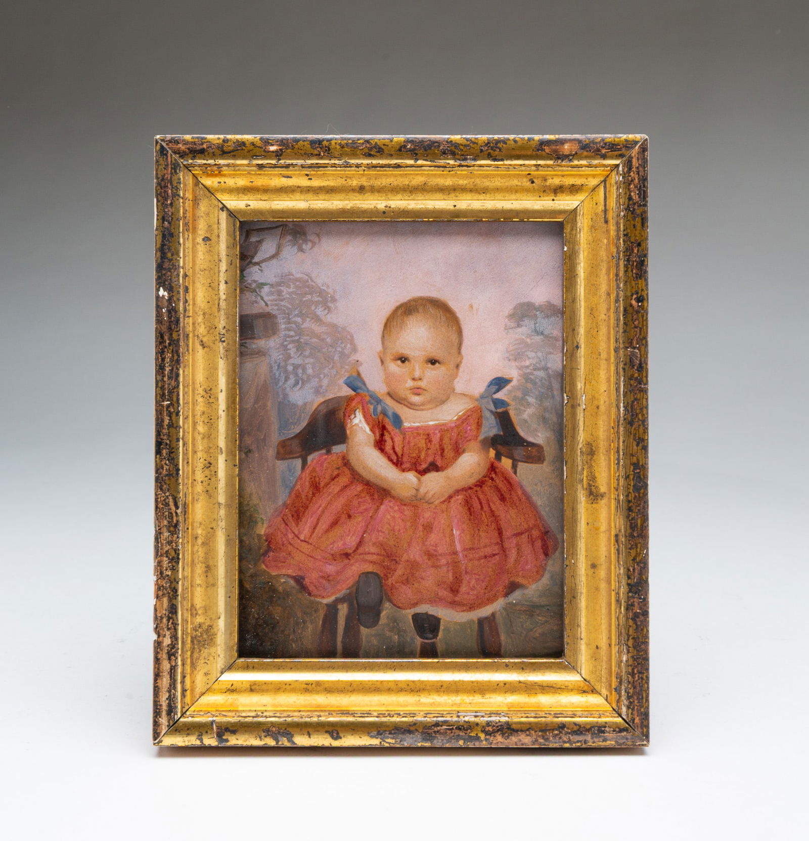 MINIATURE AMERICAN SCHOOL FOLK ART GOUACHE PORTRAIT OF A YOUNG CHILD.: 19th Century. The child wearing a pink and yellow dress with blue bows and black shoes, seated in a chair with trees in the background, in a giltwood frame. Frame, 7"h. 5.75"w.Provenance: From the