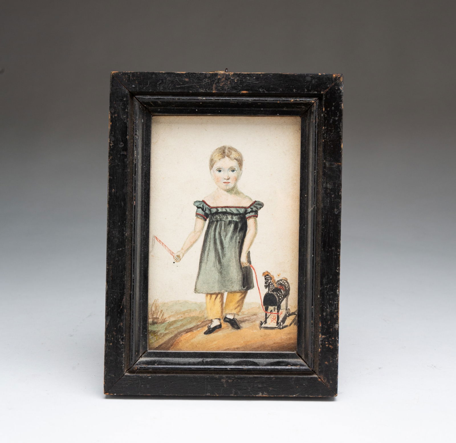 MINIATURE AMERICAN SCHOOL FOLK ART WATERCOLOR PORTRAIT OF A BOY WITH PULL TOY.: 19th Century. Full-length portrait of a boy with short sandy blonde hair and a rosy complexion wearing a blue-grey dress, yellow pants, and black shoes, pulling a horse toy with his left hand and hold