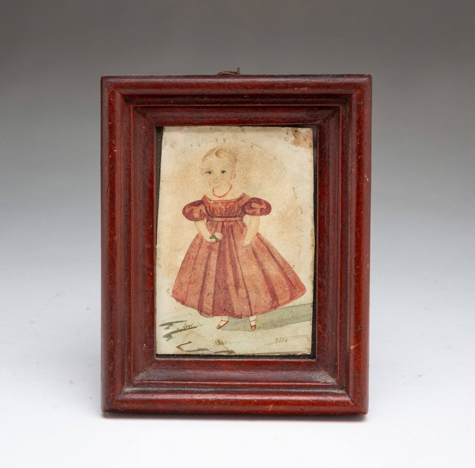 MINIATURE AMERICAN SCHOOL FOLK ART WATERCOLOR PORTRAIT OF A CHILD.: Mid-19th Century. On paper. A full-length portrait of a child with blond ringlets and dark eyes wearing a red necklace, red dress, and red shoes, holding a flower in their right hand, in an old