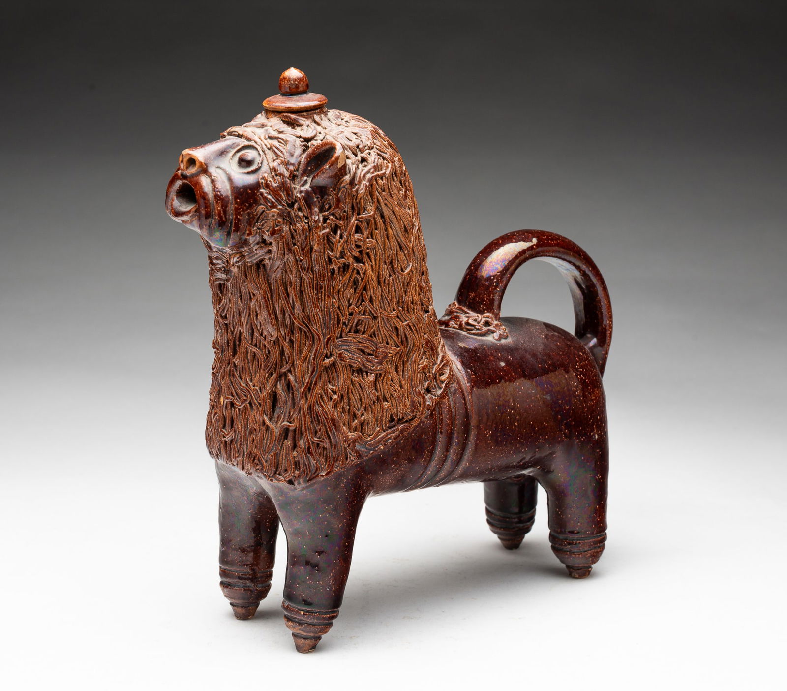 AMERICAN GLAZED REDWARE LION PITCHER.: Ca. 1900. Standing lion sculpted with lidded opening on top of head, perky ears, wide eyes, whiskers, open mouth, spaghetti mane, arched tail, and cylindrical legs with peg feet. 9.75"h. 3.25"w.