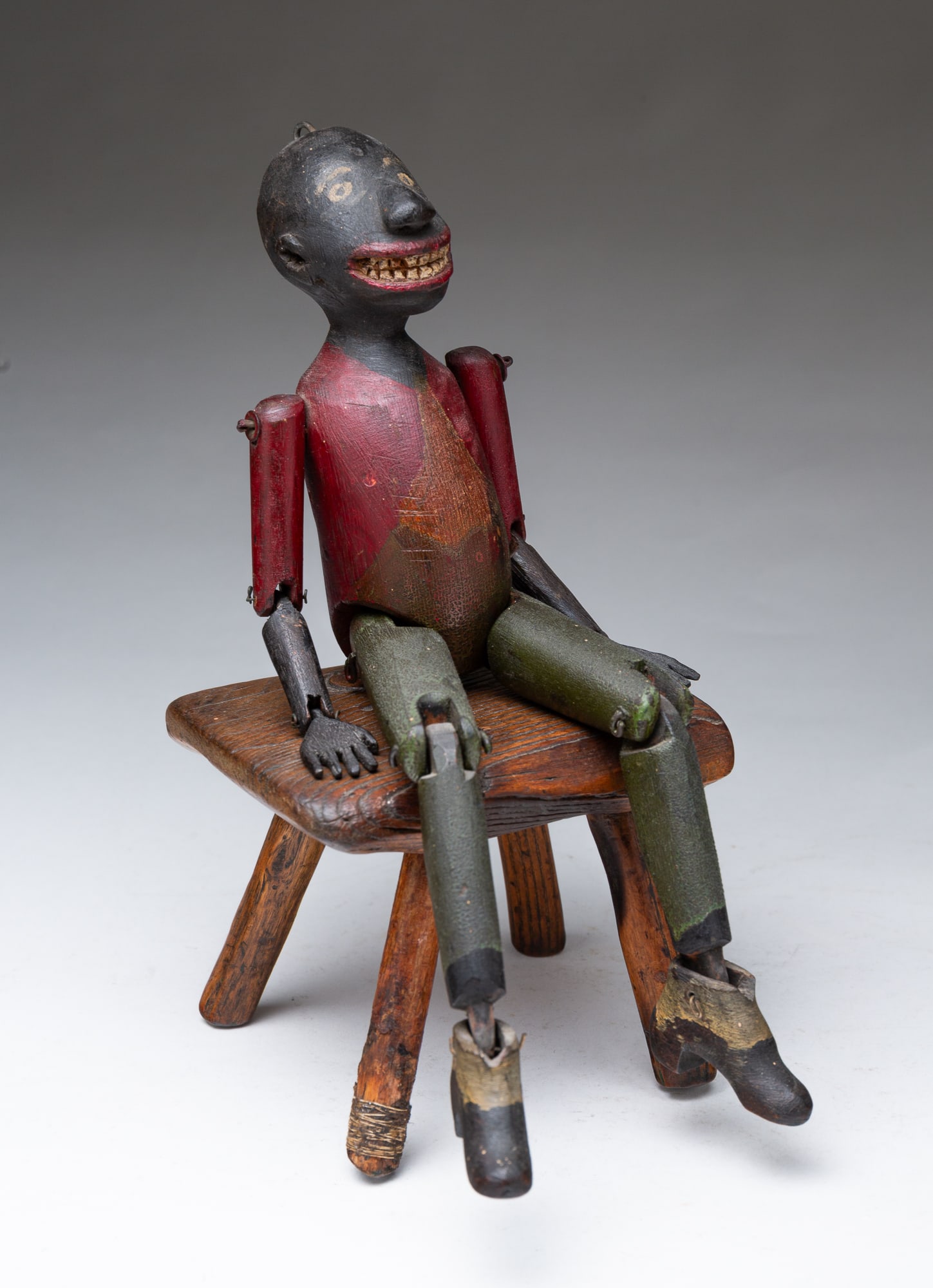 AMERICAN FOLK ART CARVED, PAINTED, AND JOINTED MAN WITH STOOL.: 19th Century. A bald male figure with carved and painted facial features, wearing a red painted coat, green painted slacks, black and white shoes and jointed limbs. Sitting upon a diminutive