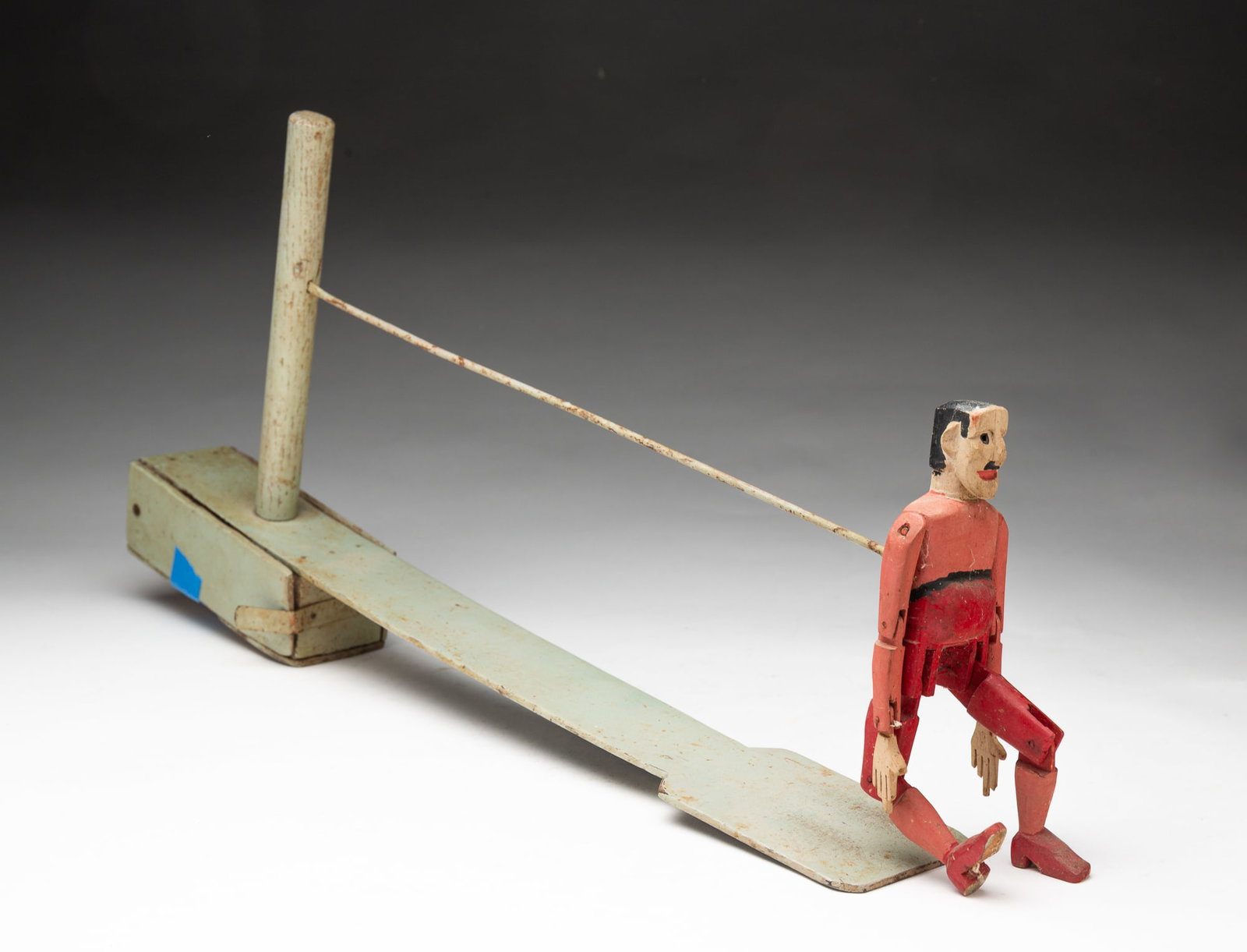 AMERICAN FOLK ART CARVED AND PAINT-DECORATED DANCING LIMBERJACK TOY.: Early-mid 20th Century. Wood, Masonite, and metal. Depicting a man with articulated joints and carved and painted facial features attached to a long, slender green-painted platform. 9"h. 2.5"w.