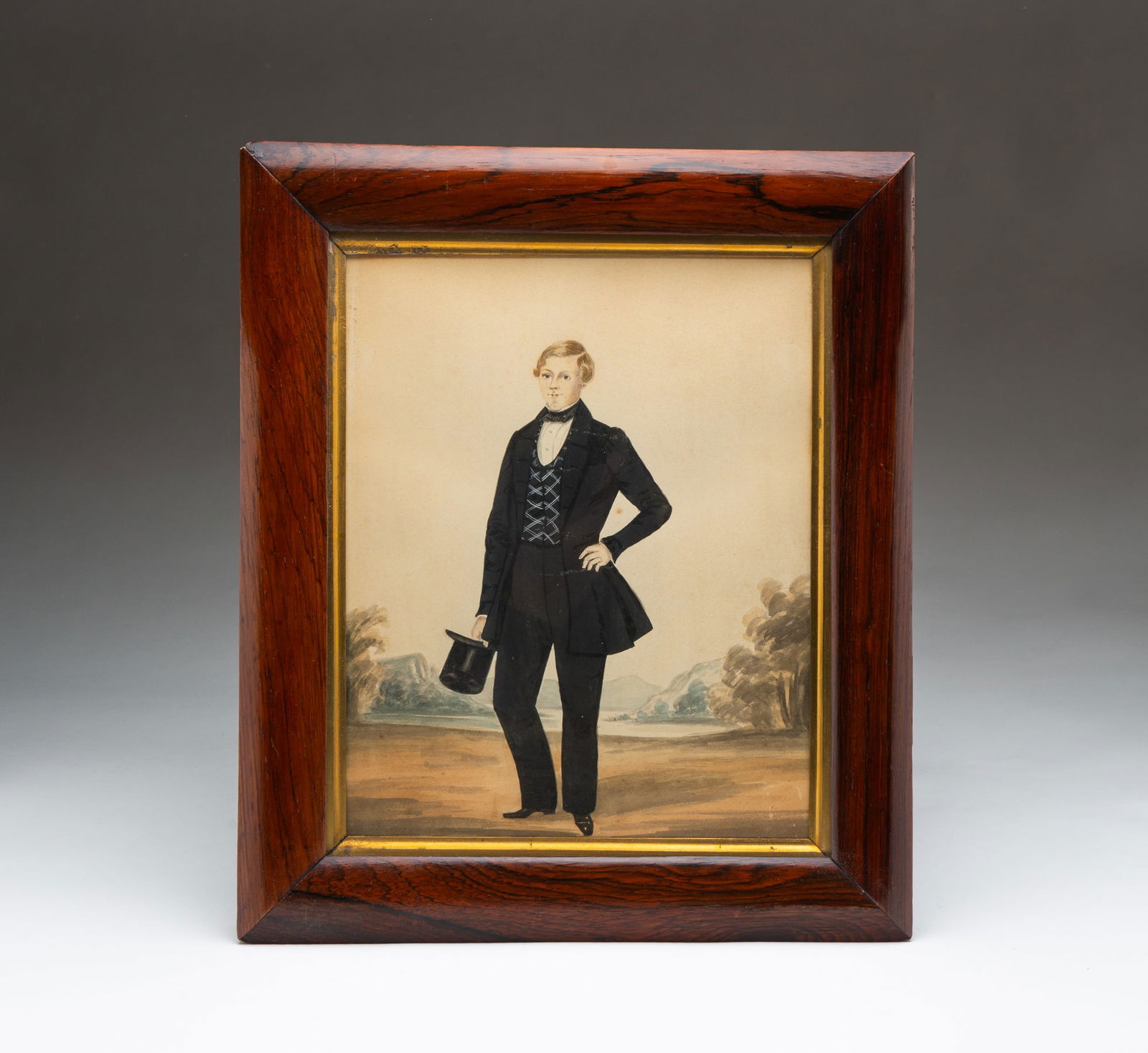 AMERICAN SCHOOL FOLK ART WATERCOLOR PORTRAIT OF A GENTLEMAN.: Mid-19th Century. On paper. A full-length depiction of a man looking straight ahead, wearing a black coat with a striped vest, white shirt, black necktie, and holding his hat standing in front of a se