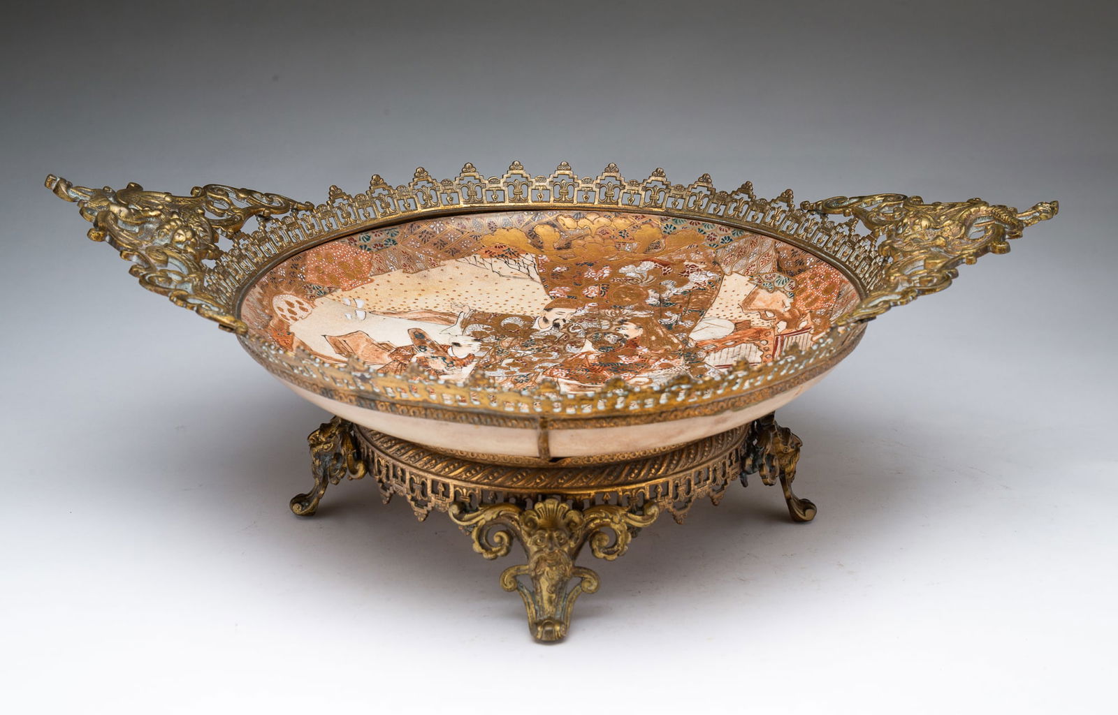 JAPANESE SATSUMA BOWL IN ORNATE BRASS ORMOLU FRAME.: Late 19th Century. Bowl has scene of two men and a young boy in ornate robes, mountains and sea in background, multi colored border and fine gold trim. In a reticulated frame with serpent handles and