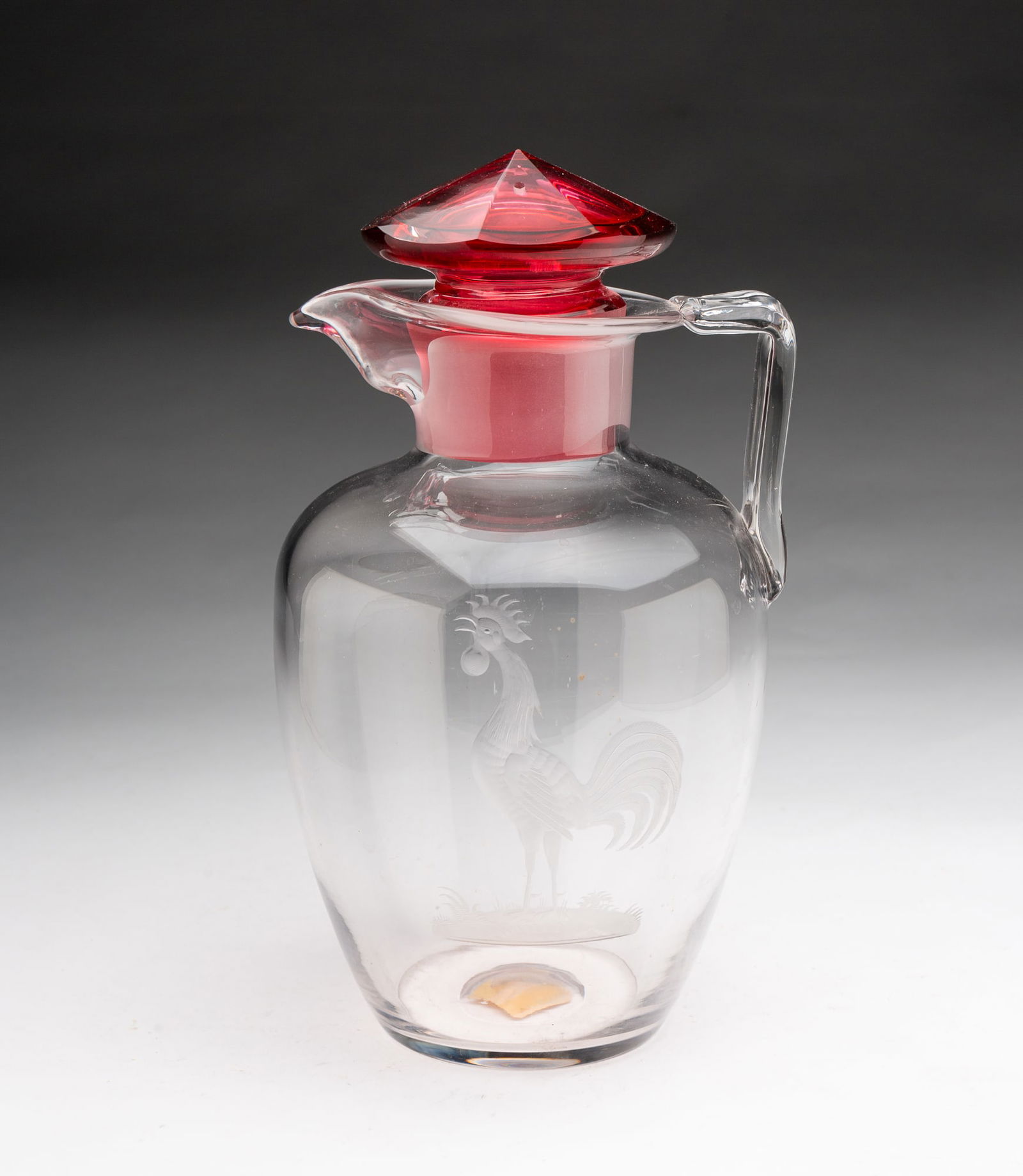STEUBEN (CORNING, NEW YORK) ROOSTER-ETCHED CLEAR GLASS COCKTAIL PITCHER WITH CRANBERRY STOPPER.: American, First quarter 20th Century. Shape 7056. Clear pitcher with etched rooster, applied handle, and faceted cranberry stopper with keyhole notch. Signed with acid etched fleur-de-lis mark.