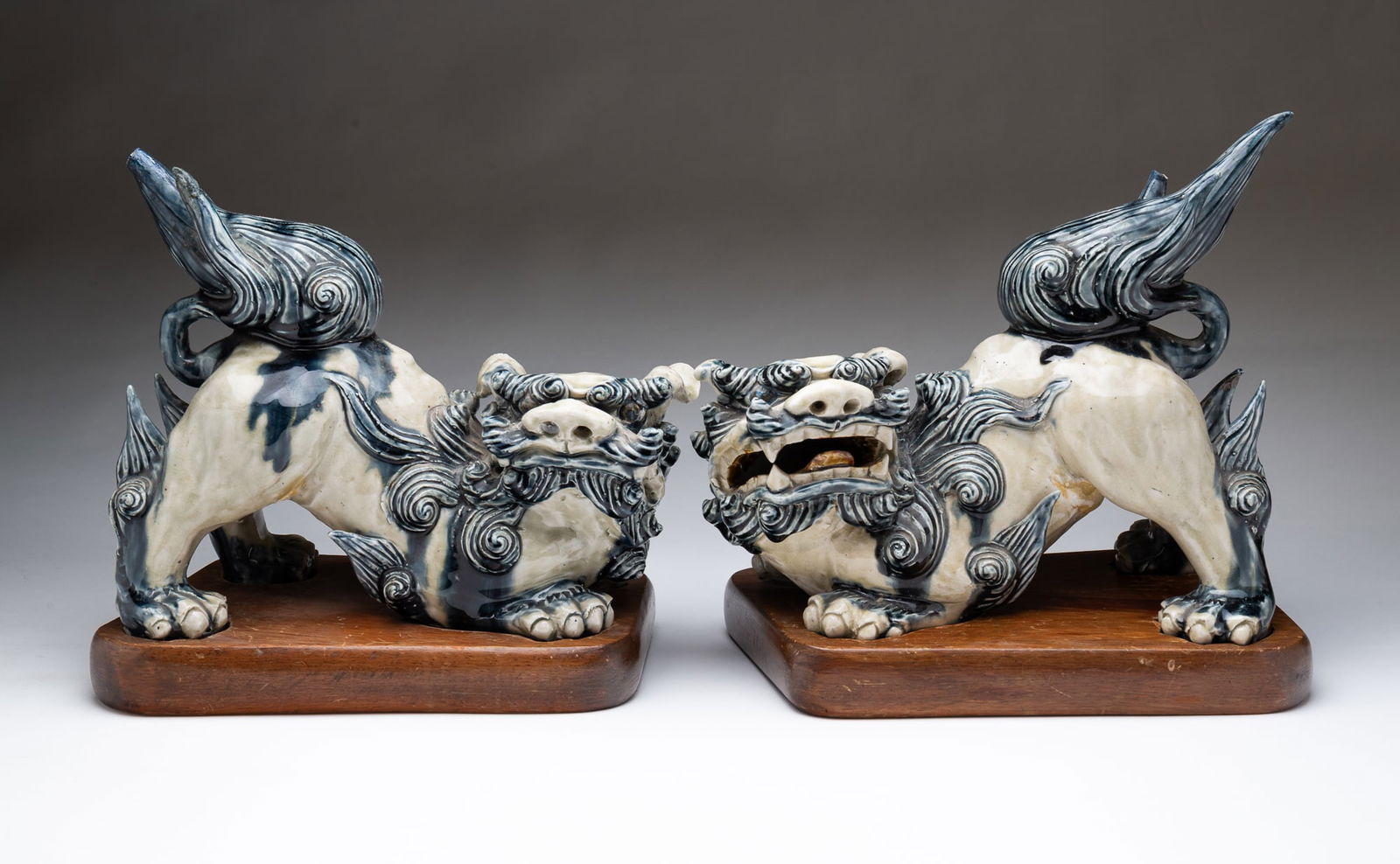 PAIR OF CHINESE GLAZED PORCELAIN FOO DOGS.: Mid-20th Century. Opposing foo dogs, each in a playful crouching pose, the right one with an open mouth, each glazed in blue and white with spiraling clouds, with fitted wooden stands. 11.5"h. 12"w.
