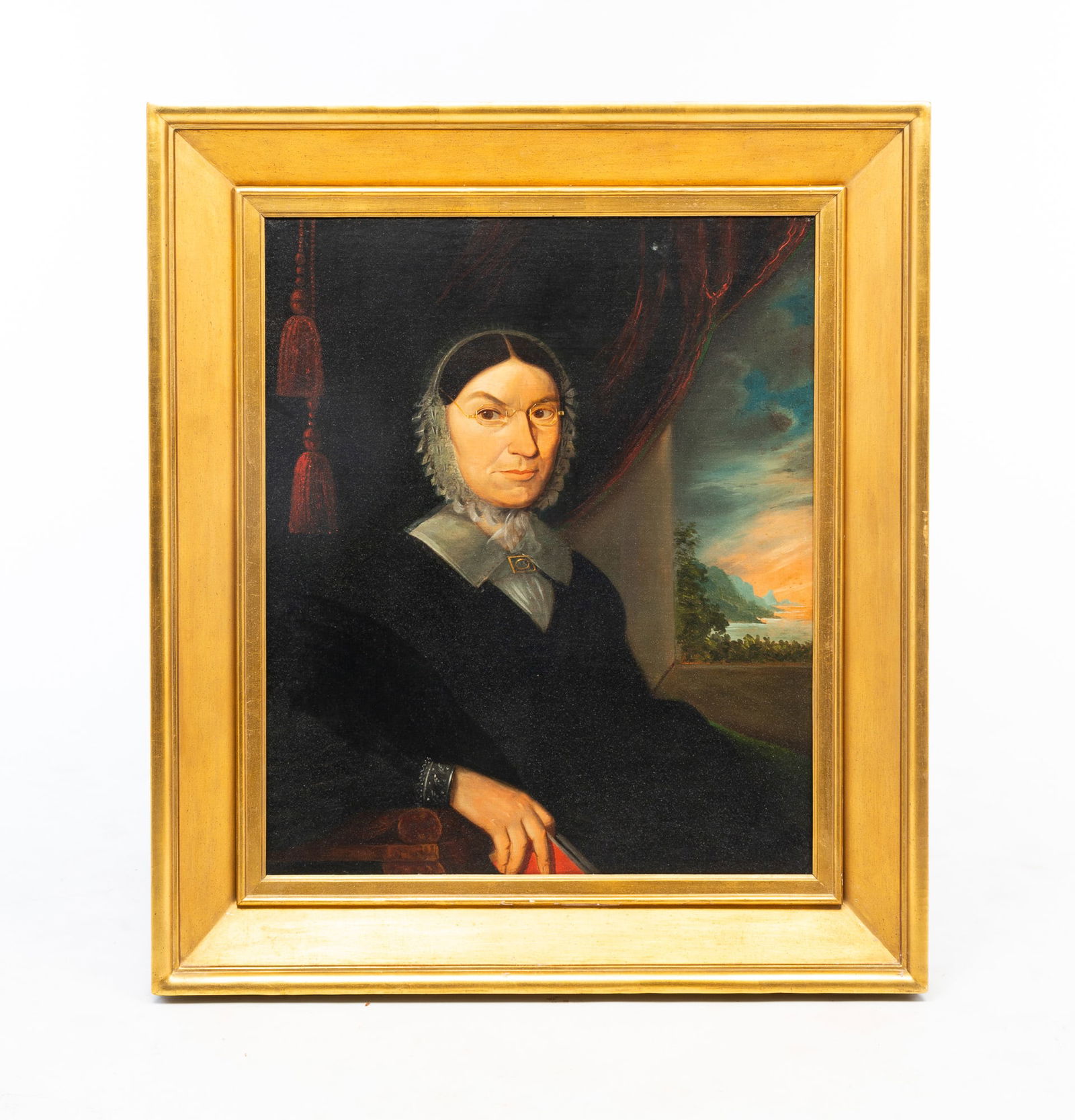 LARGE AMERICAN SCHOOL FOLK ART OIL ON BOARD PORTRAIT OF A LADY WEARING A BONNET.: American, ca. 1830. Oil on board. Portrait of a lady wearing a white bonnet, glasses, white collar, gold brooch, and a black dress, casually holding a book in her right hand, seated before heavy red d