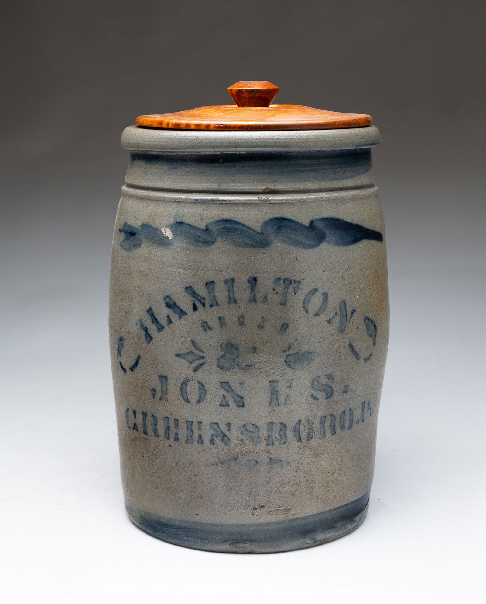 HAMILTON AND JONES (GREENSBORO, PENNSYLVANIA) COBALT-DECORATED STONEWARE JAR. 9.5"h. 6.5"d.: American, Second half 19th Century. Well formed lip, tooled band, stenciled "HAMILTON JONES, GREENSBORO, PA", freehand cobalt stripes and a squiggle. Includes a contemporary turned tiger maple lid.SHI
