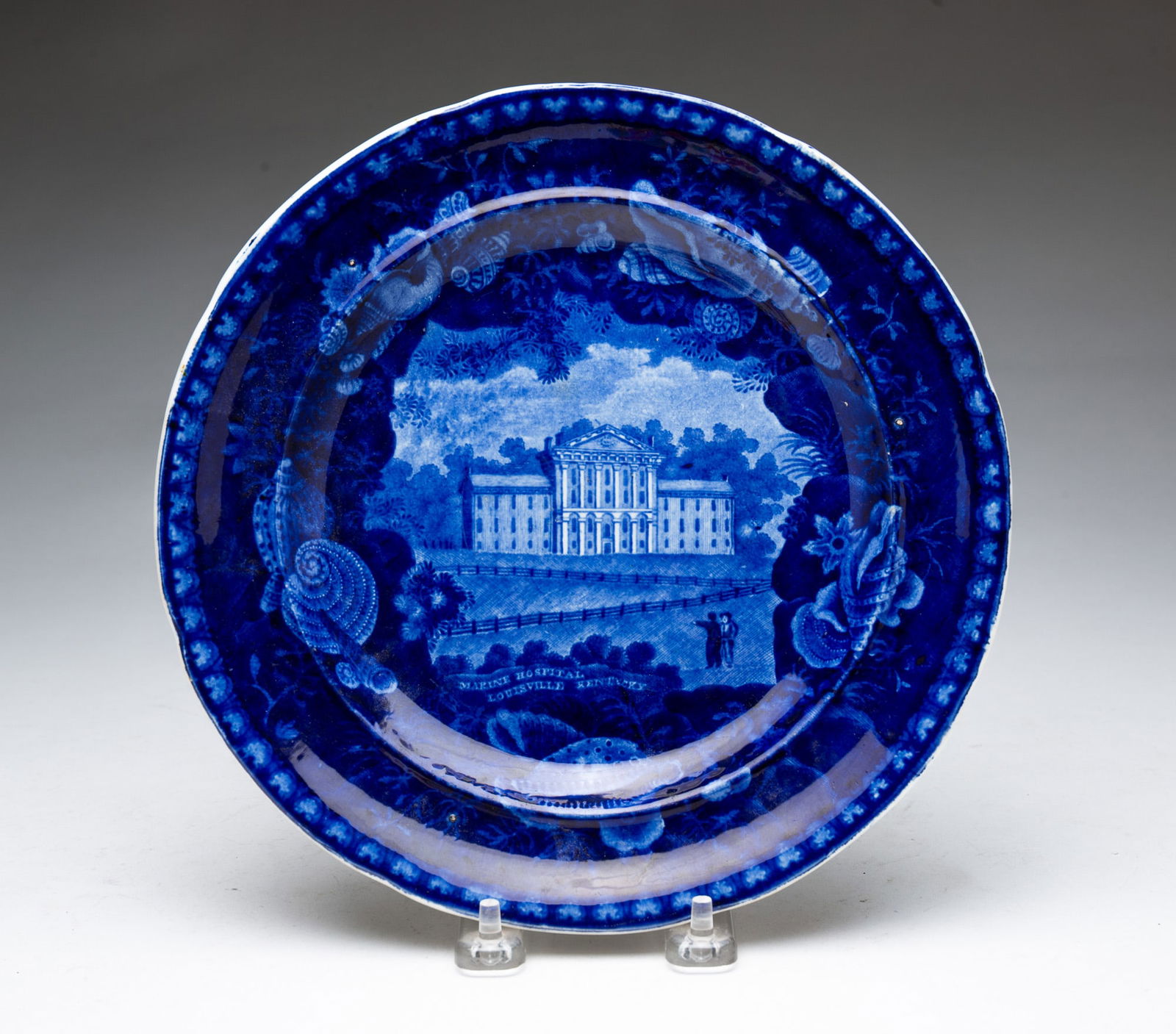 ENOCH WOOD HISTORICAL "BLUE MARINE HOSPITAL, LOUISVILLE, KENTUCKY" PLATE. 9"d.: English, Mid-19th Century. Shell border with the hospital at the center. Impressed mark on reverse.SHIPPING: All purchases from Amelia Jeffers, Auctioneers & Appraisers may be picked up by you or your
