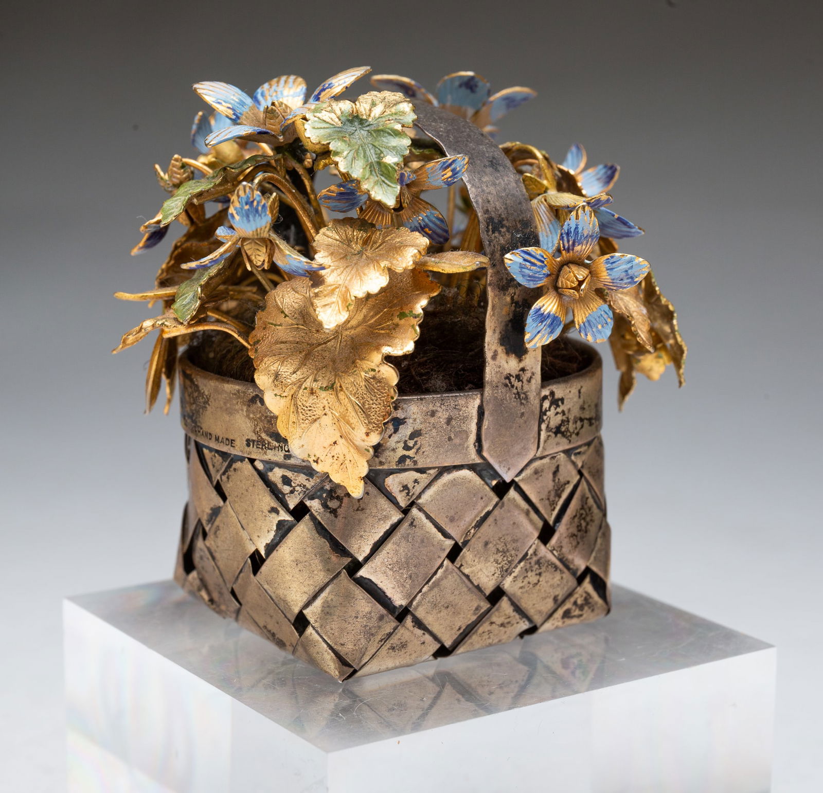 CARTIER (NEW YORK) GILT AND ENAMEL FLOWERS AND LEAVES IN A STERLING SILVER BASKET. 3.25"h. 3.25"w.: American, ca. 1950. Gilt and enameled blue flowers and green enameled leaves contained in a Cartier sterling silver woven basket with handle. Stamped "Cartier Hand Made Sterling." Total weight, 5.28 o