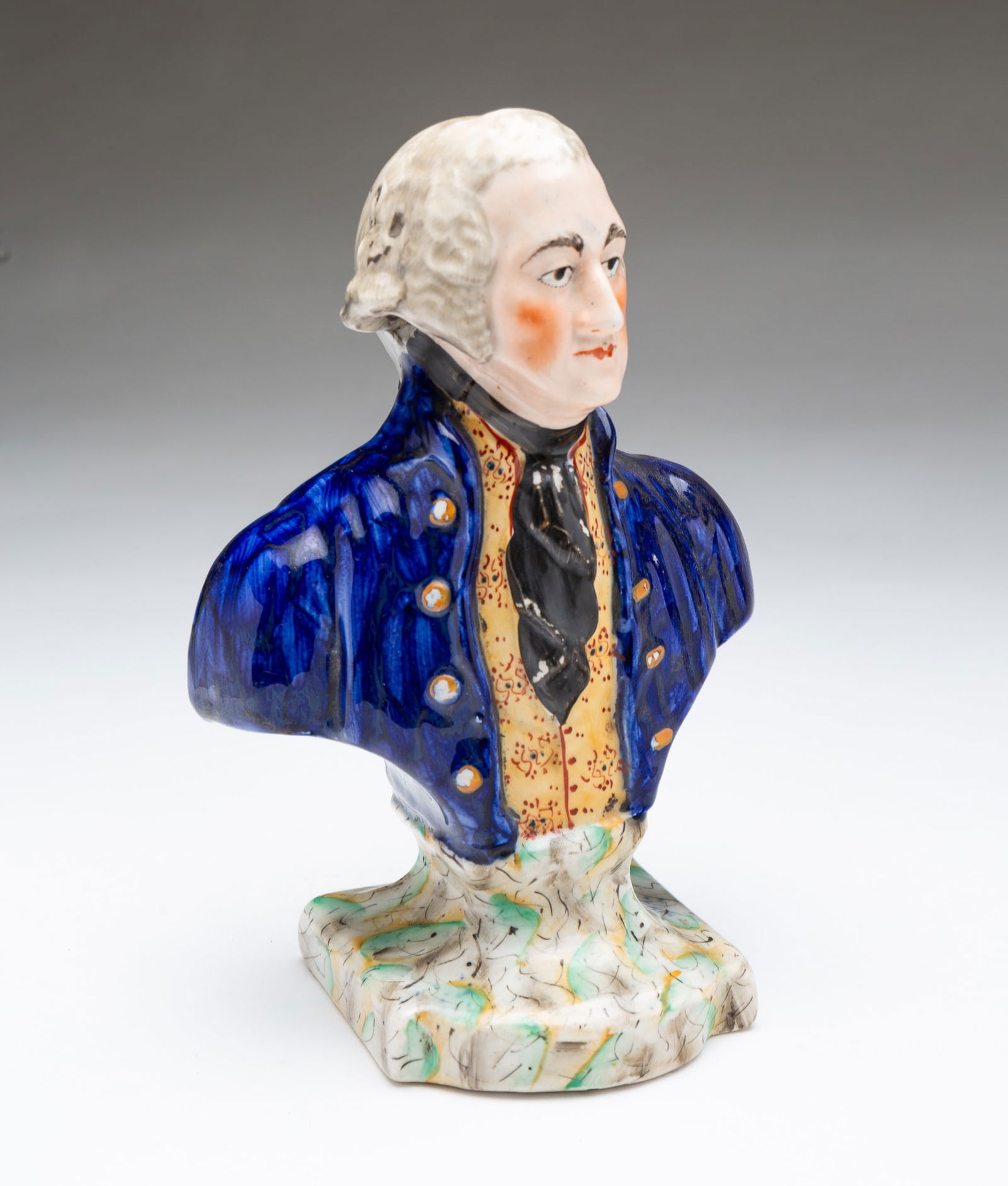 STAFFORDSHIRE BUST OF GEORGE WASHINGTON. 7.75"h.: English, 19th Century. He is portrayed with coiffed gray hair, painted face, wearing a blue jacket with gold buttons, a yellow vest with pattern, and a black cravat on a plinth colored in green, yello