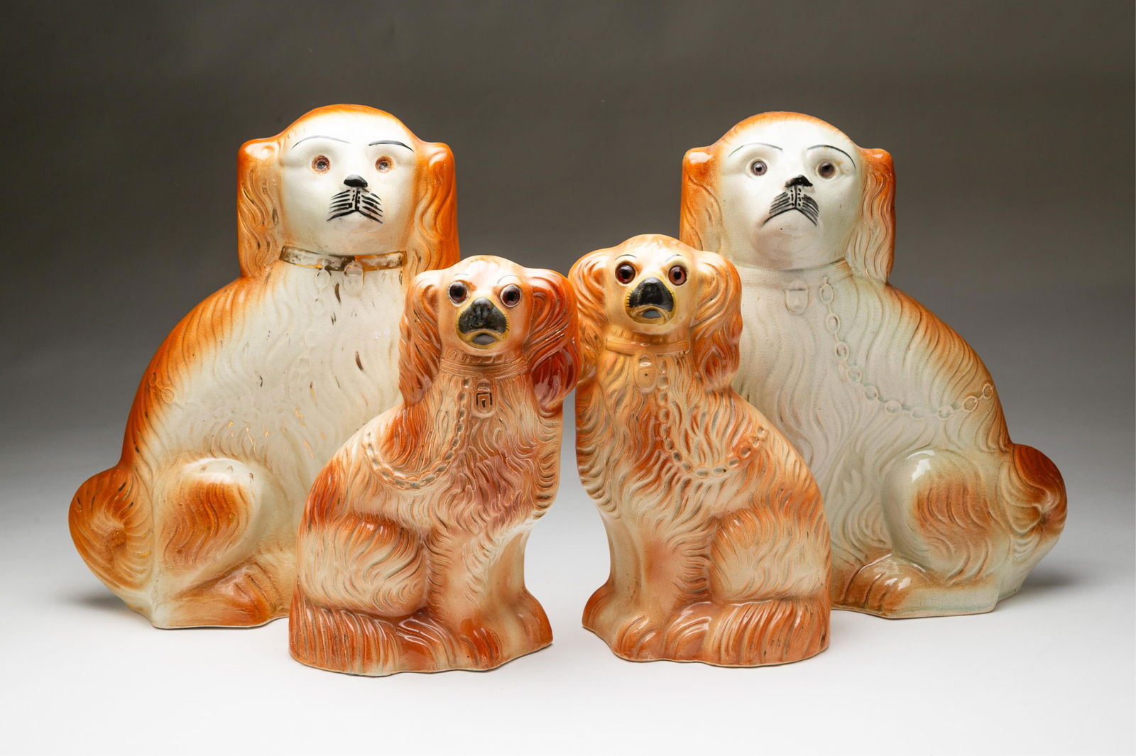 TWO PAIRS OF STAFFORDSHIRE SPANIELS WITH GLASS EYES. The larger pair, 14"h. 11"w.: English, Second-half 19th Century. Both pairs cream and orange. The larger pair with luster gilt accents, one has a purple glass eye and an amber glass eye, the other without eyes. The smaller pair wi