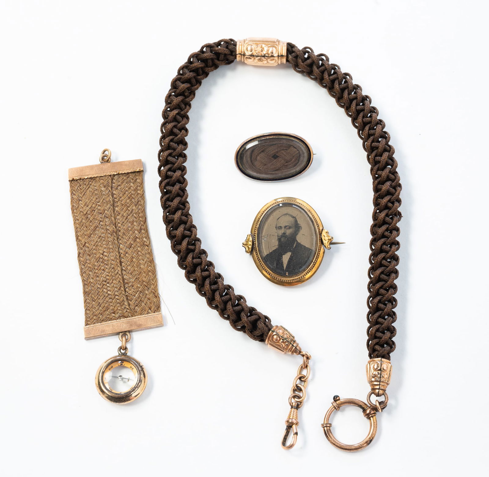 FOUR VICTORIAN WOVEN HAIR-WORK MOURNING JEWELRY ITEMS. Necklace, 15"l.: American, Second half 19th Century. Unmarked. Two gold-filled or plated brooches; one with braided hair and the other with a portrait and braided hair, a braided hair fob with a compass, and a