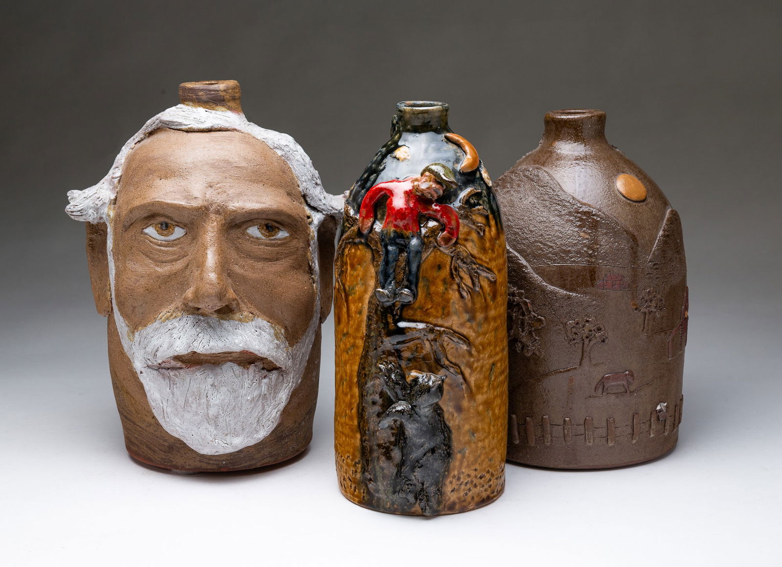 THREE TURKEY MT. POTTERY CERAMICS BY TOM AND JUDY TOUCHSTONE. Largest, 11"h.: Armuchee, Georgia, Contemporary. Ceramic. A glazed jug with a bear chasing a man up a tree, A decorated jug with a barnyard scene with animals, and a face jug depicting a man with white hair and beard