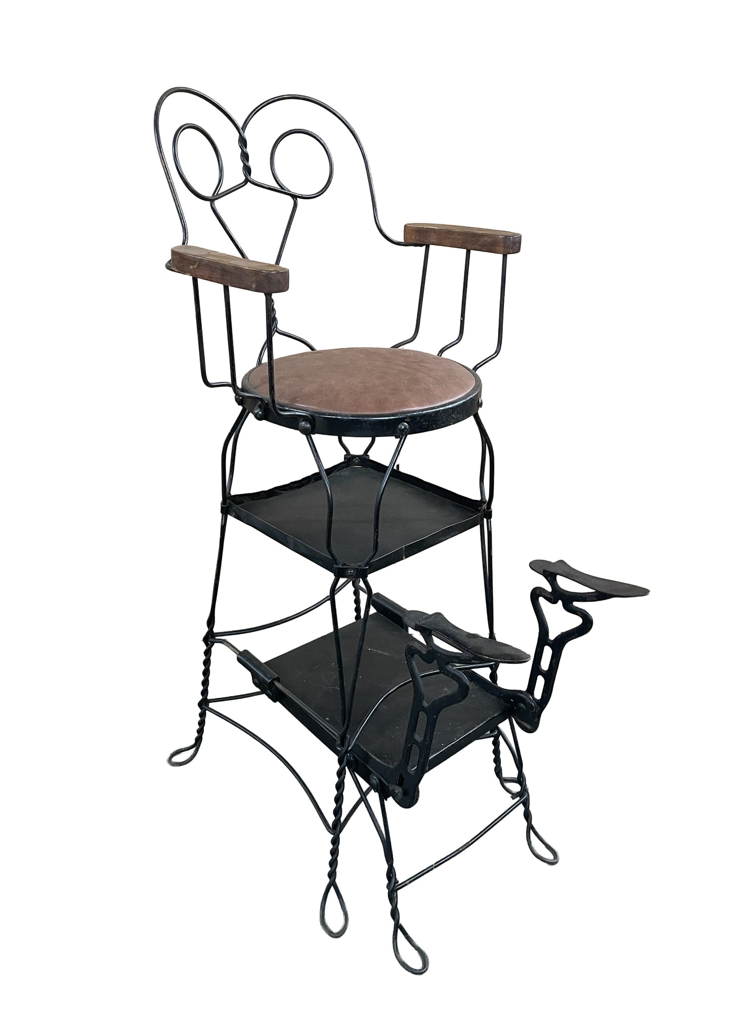 WROUGHT AND CAST IRON SHOESHINE CHAIR.: American, ca. 1900. Wrought iron heart-shaped back, wooden armrests, padded brown seat, and extendable cast iron shoe rests on bottom. 52.5"h. 36" seat h. 22.5"w. 30"d. (not extended)Provenance: From