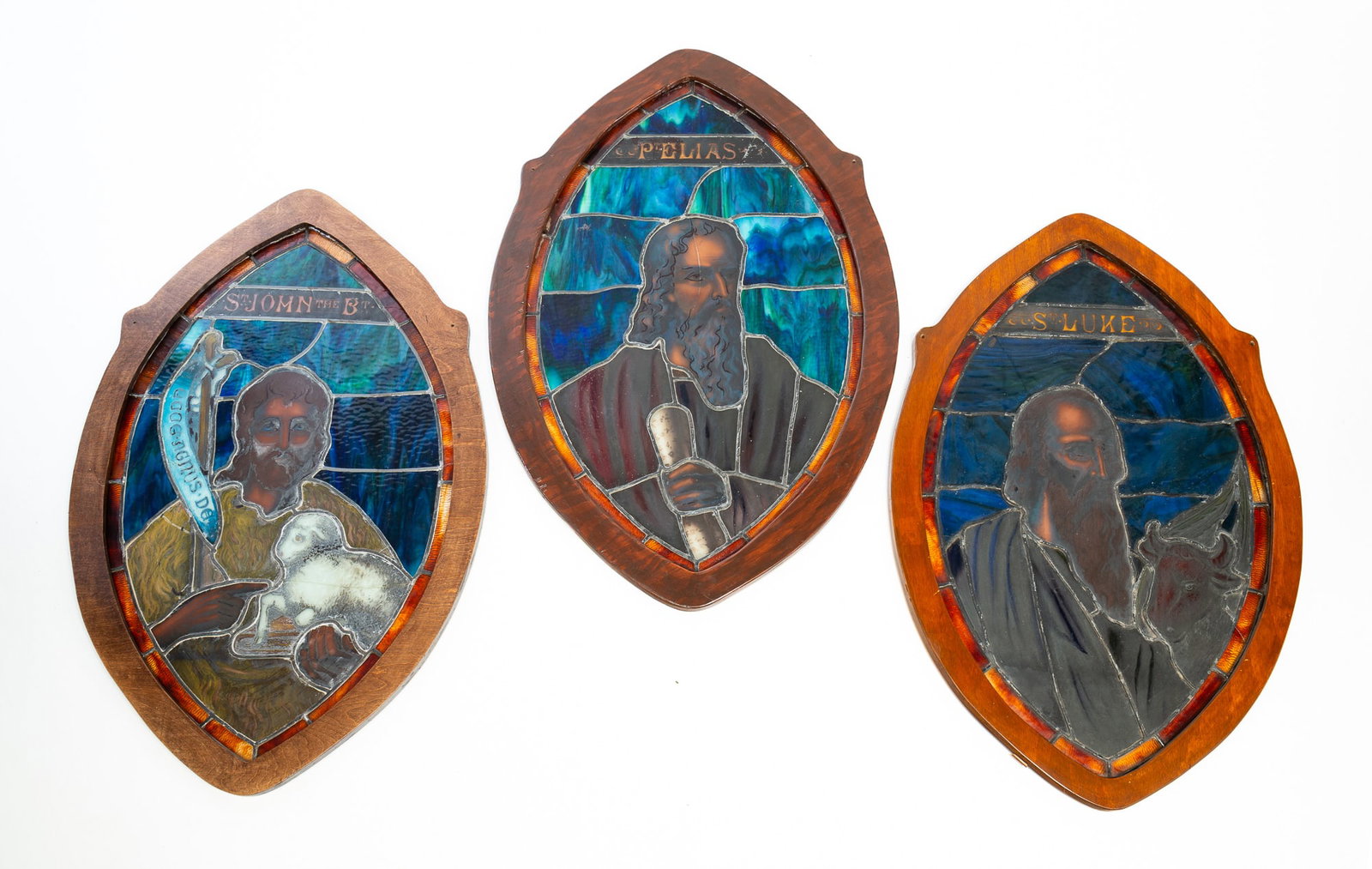 THREE AMERICAN STAINED AND LEADED GLASS CHURCH WINDOWS.: Second half 19th Century. Plywood frames. Ellipse with pointed ends, amber borders, blue slag glass backgrounds, and stained glass saints. "John the Baptist", "Prophet Elias" and "Saint Luke". 38.5"h.
