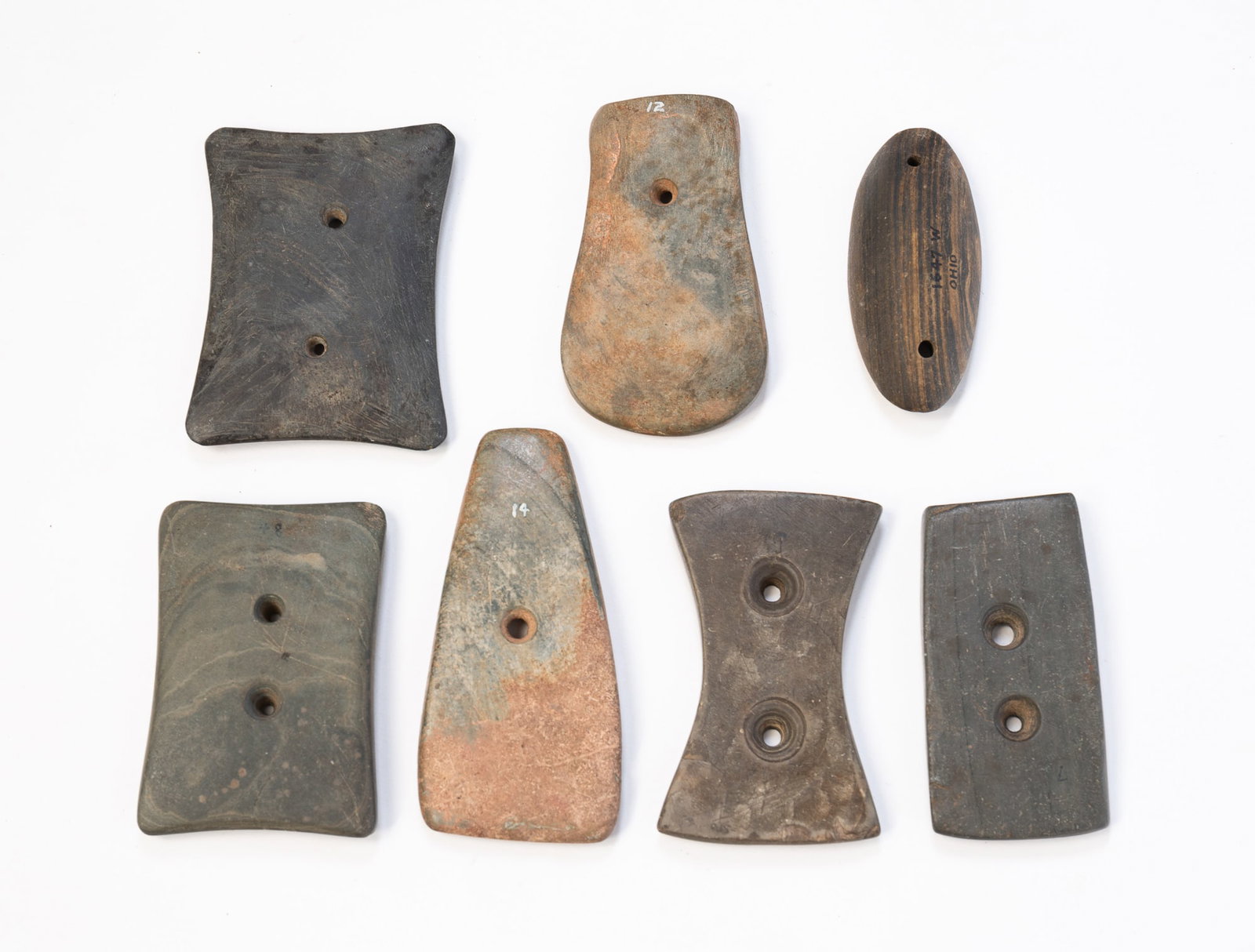 SEVEN NATIVE AMERICAN STONE GORGETS.: SEVEN NATIVE AMERICAN STONE GORGETS. Four are grey stone with double holes, two are pink and grey with single holes, and one is black and tan with double holes, and identified as found in Ohio. From