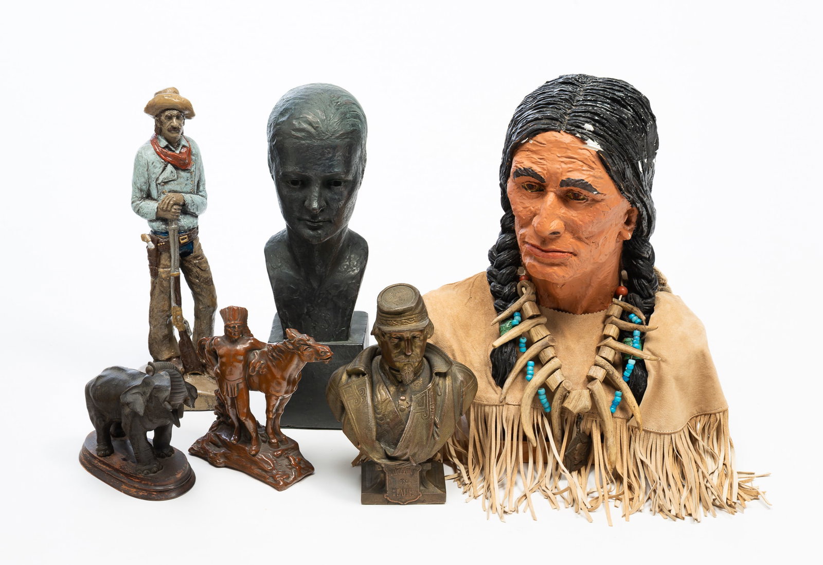 SIX FIGURAL BUSTS, STATUES, AND FIGURINES.: SIX FIGURAL BUSTS, STATUES, AND FIGURINES. American, 20th Century. A large realistic plaster bust of a Native American wearing buckskin shirt, a bone and antler necklace, mounted on a wooden board,