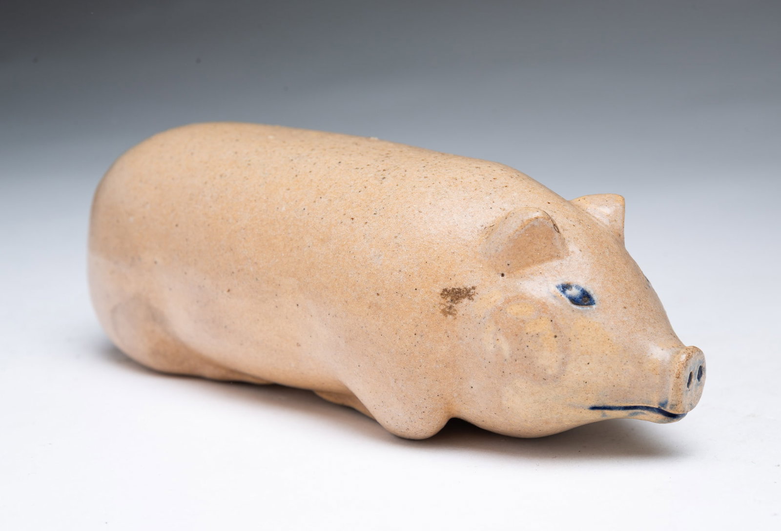 COBALT-DECORATED STONEWARE PIG FLASK.: COBALT-DECORATED STONEWARE PIG FLASK. American, Late 19th Century. Reclining pig with perky ears and legs tucked underneath, having painted cobalt eyes, snout, and mouth, with a hole in the rear for a