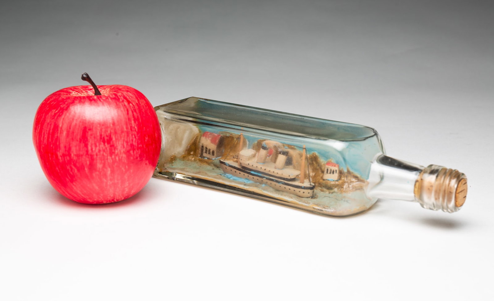 MINIATURE FOLK ART SHIP IN A BOTTLE.: MINIATURE FOLK ART SHIP IN A BOTTLE. American, 20th Century. A steamer ship painted in white with black trim traveling on the sea past a mountainside village, in a squared glass bottle with cork