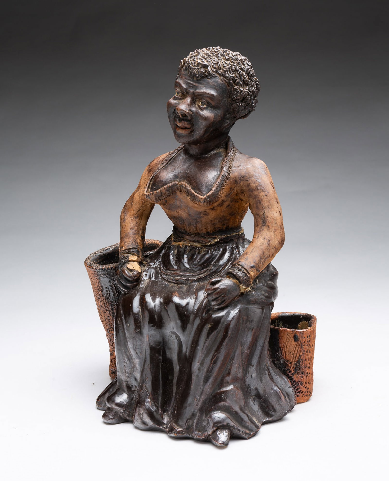 ALBANY SLIP GLAZED TERRACOTTA BLACK AMERICANA FIGURE OF A SEATED WOMAN BETWEEN OPEN BASKETS.: ALBANY SLIP GLAZED TERRACOTTA BLACK AMERICANA FIGURE OF A SEATED WOMAN BETWEEN OPEN BASKETS. American, 19th Century. Sculpted figure of a woman wearing a low-cut dress, seated on a tree stump between
