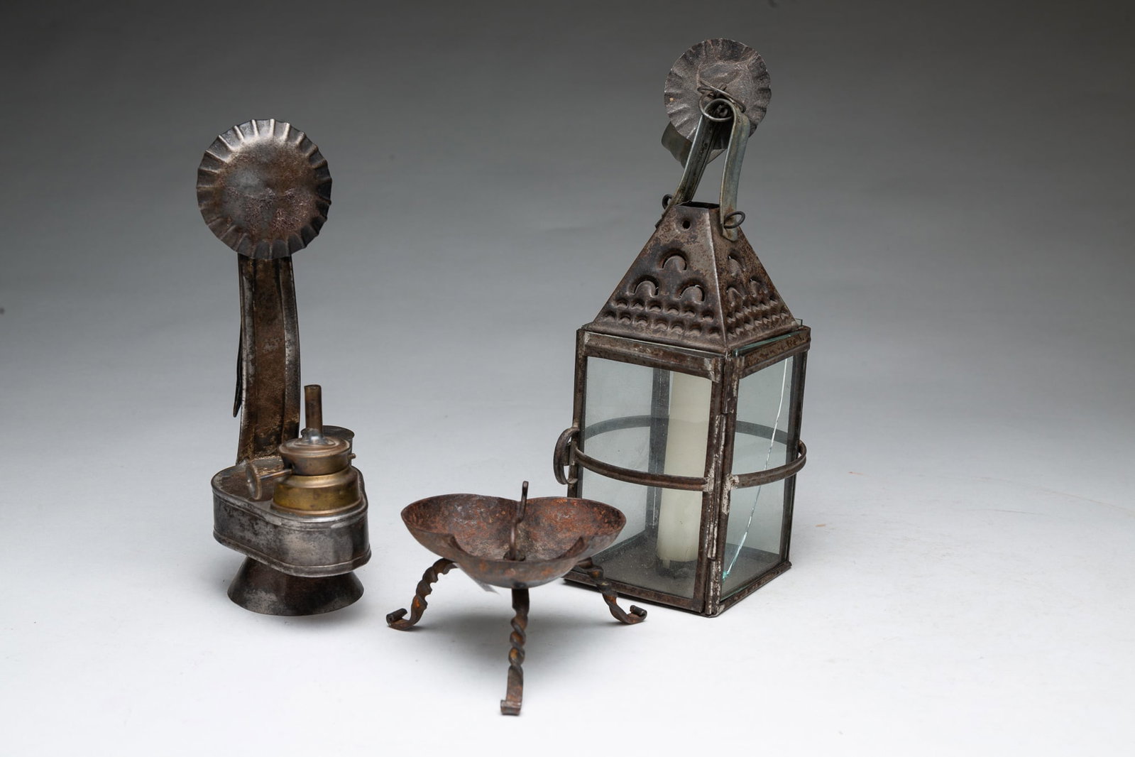 THREE LIGHTING DEVICES.: THREE LIGHTING DEVICES. American, 19th Century. A footed clover-shaped iron grease lamp. a fluid burning lamp with a reflector, and a glass pane candle lantern with a ring handle. Tallest, 11.75"h. Pr
