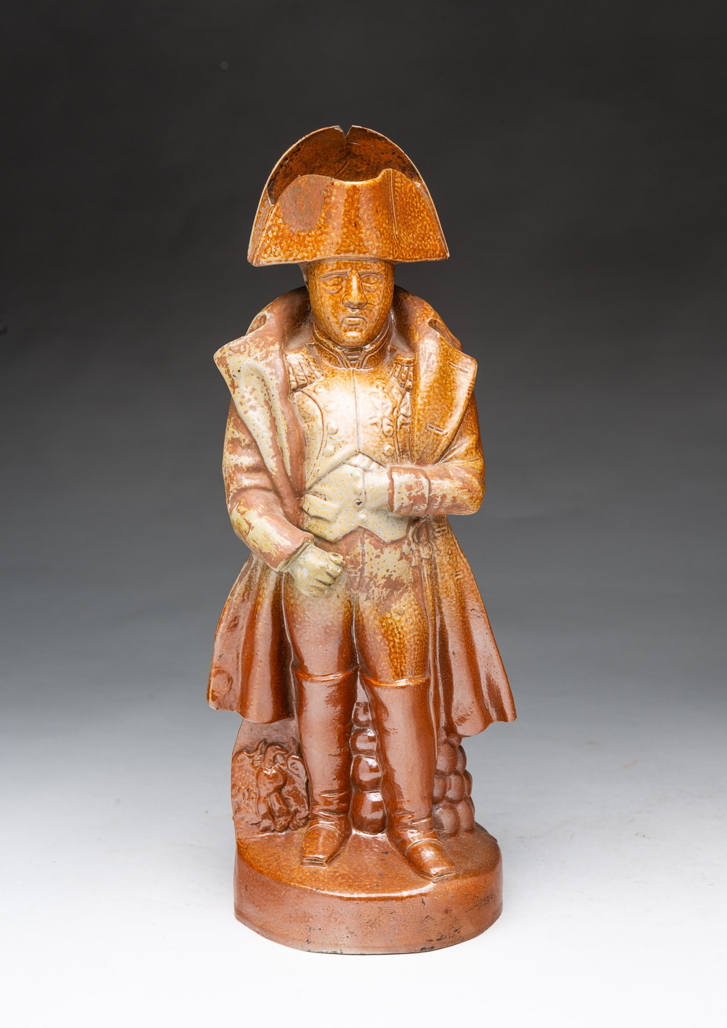 GLAZED STONEWARE PITCHER IN THE FORM OF NAPOLEON BONAPARTE.: GLAZED STONEWARE PITCHER IN THE FORM OF NAPOLEON BONAPARTE. American, Mid-19th Century. Salt-glazed stoneware pitcher in mottled brown and tan glaze, of a standing Napoleon wearing his large hat, an o