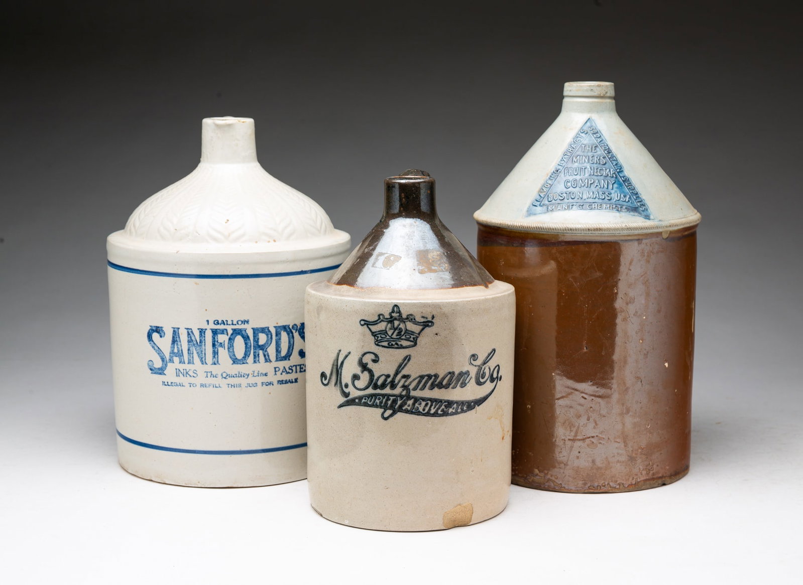 THREE STONEWARE ADVERTISING JUGS.: THREE STONEWARE ADVERTISING JUGS. American, Late 19th and early 20th Century. One is brown with white conical top with embossed label in a blue triangle "Miner Fruit Nectar Company, Boston, Mass,