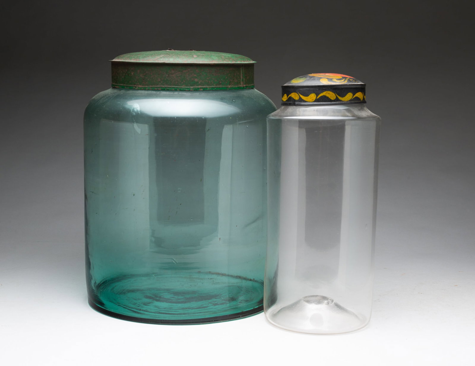 TWO BLOWN GLASS STORAGE JARS WITH LIDS, INCL. TOLE-LIDDED EXAMPLE.: TWO BLOWN GLASS STORAGE JARS WITH LIDS, INCL. TOLE-LIDDED EXAMPLE. American, 19th Century. Both of cylindrical form. Largest in bottle glass with green painted lid, 13"h., and the other in clear