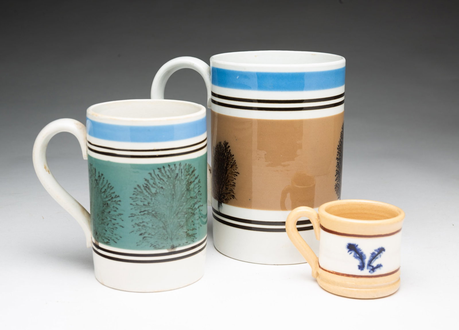 THREE CONTEMPORARY MOCHAWARE DECORATED ITEMS.: THREE CONTEMPORARY MOCHAWARE DECORATED ITEMS. American, 20th Century. A large mug with white ground, tan and blue bands, and seaweed decoration, 6.5"h., a mug with white ground, black, green, and