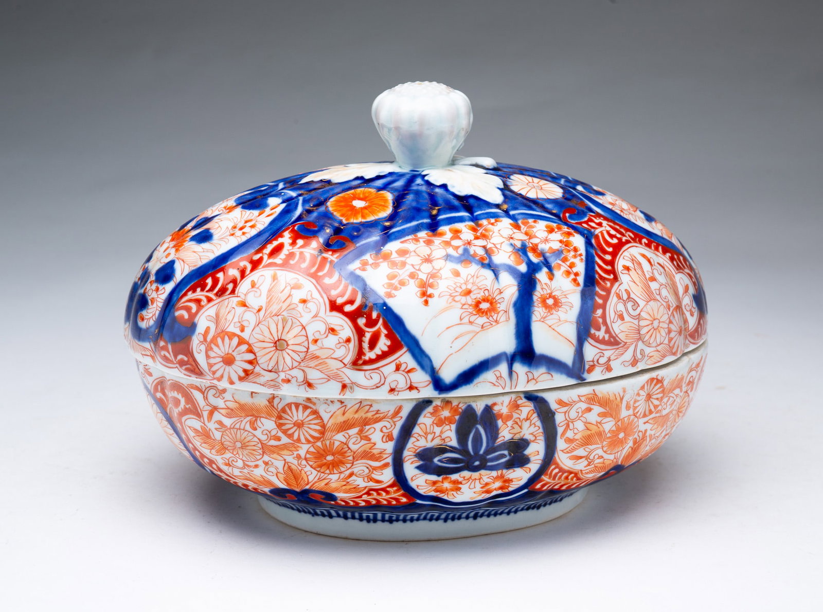 JAPANESE IMARI PORCELAIN COVERED TUREEN.: JAPANESE IMARI PORCELAIN COVERED TUREEN. Japan, 19th Century. Of oval form with molded lobes, the lid having a fruit-form finial, decorated all over in orange and blue floral and foliate motifs with