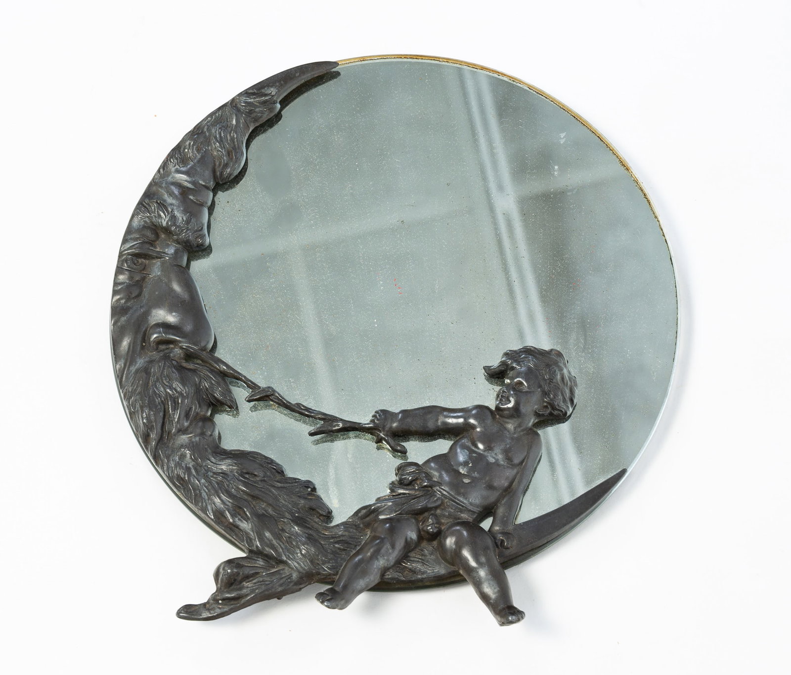PATINATED BRONZE "MAN IN THE MOON" MIRROR.: PATINATED BRONZE "MAN IN THE MOON" MIRROR. American, Late 19th or Early 20th Century. Circular wall mirror embellished with a patinated bronze scene of a cherub using a leafy stick to tickle the nose
