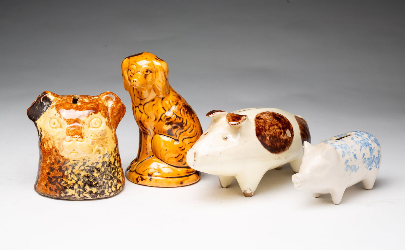 FOUR GLAZED POTTERY FIGURAL STILL BANKS, INCL. ROSEVILLE.: FOUR GLAZED POTTERY FIGURAL STILL BANKS, INCL. ROSEVILLE. American, 19th or 20th Century. Comprising two pigs, a seated spaniel, and a Roseville dog's head example. Spaniel, 5.5"h. Provenance: The