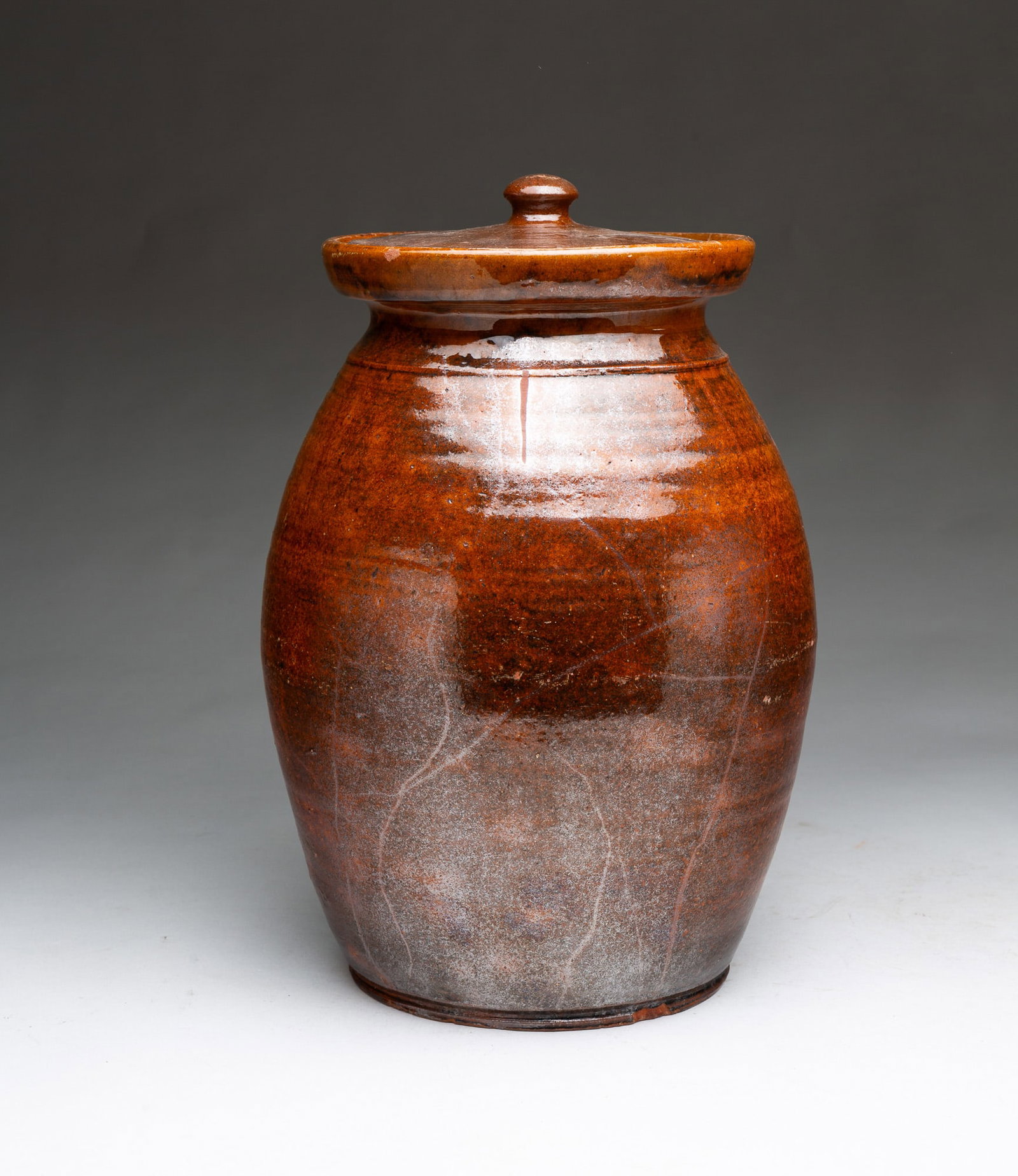 GLAZED REDWARE JAR WITH LID.: GLAZED REDWARE JAR WITH LID. American, 19th Century. Of ovoid form with flared top rim and pronounced lip and foot, lid with finial, tooled neck, and mottled orange and manganese glaze with