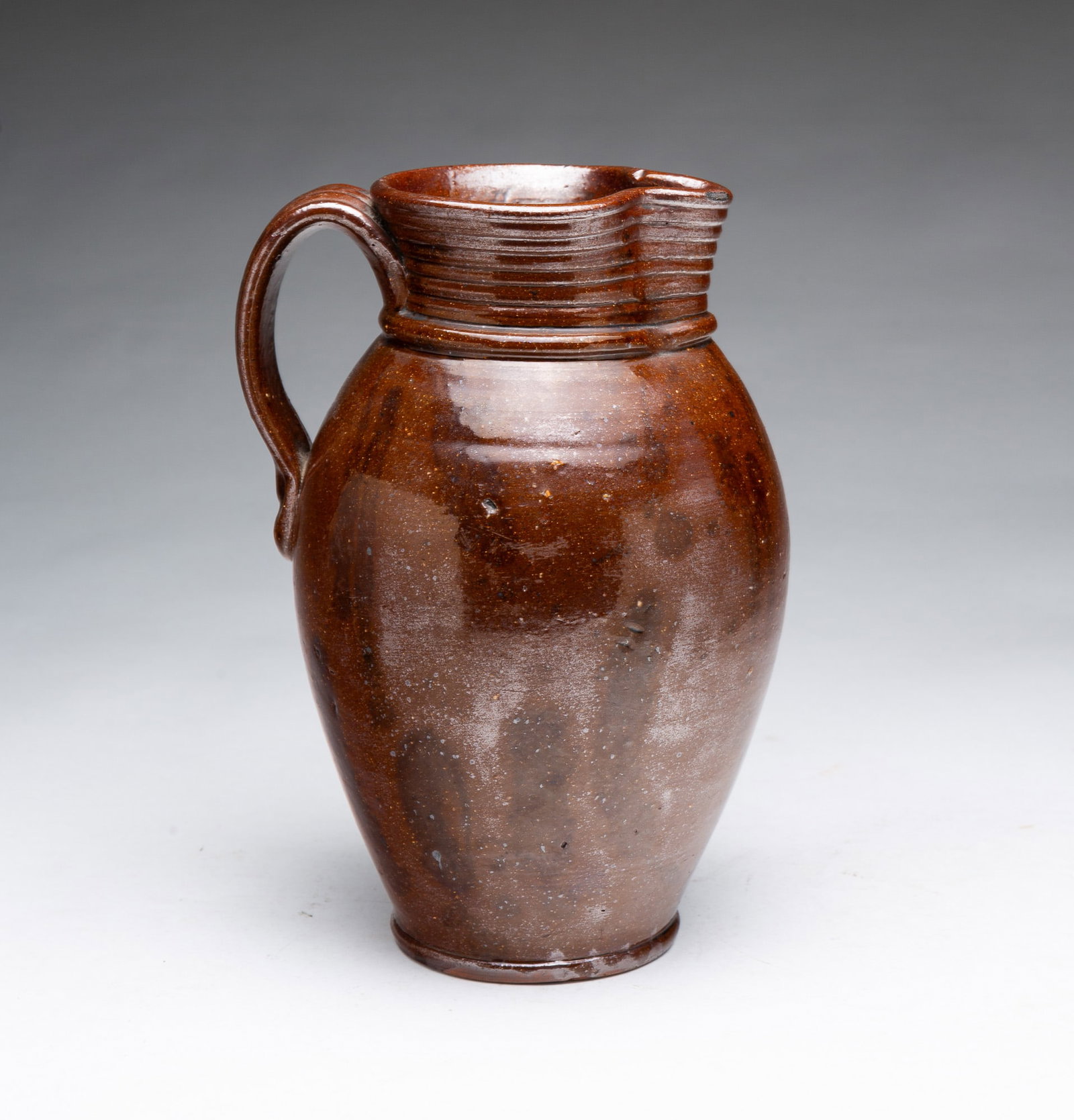 UNUSUAL FORM ALBANY SLIP GLAZED REDWARE PITCHER.: UNUSUAL FORM ALBANY SLIP GLAZED REDWARE PITCHER. American, 19th Century. Of ovoid form with flared rim, spout with incised bands, rolled collar, applied strap handle with thumbprints at terminals,