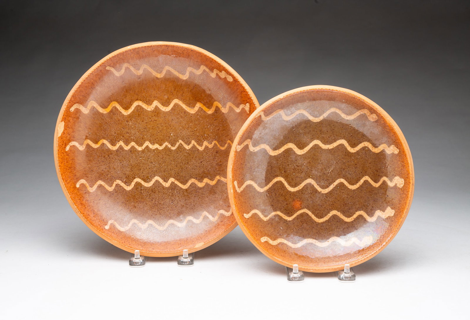 TWO SLIP-DECORATED REDWARE PLATES.: TWO SLIP-DECORATED REDWARE PLATES. American, 20th Century. Each with orange and brown mottled glaze, the center decorated with wavy lines in off-white slip, and with a coggled rim. The larger, 10"d.