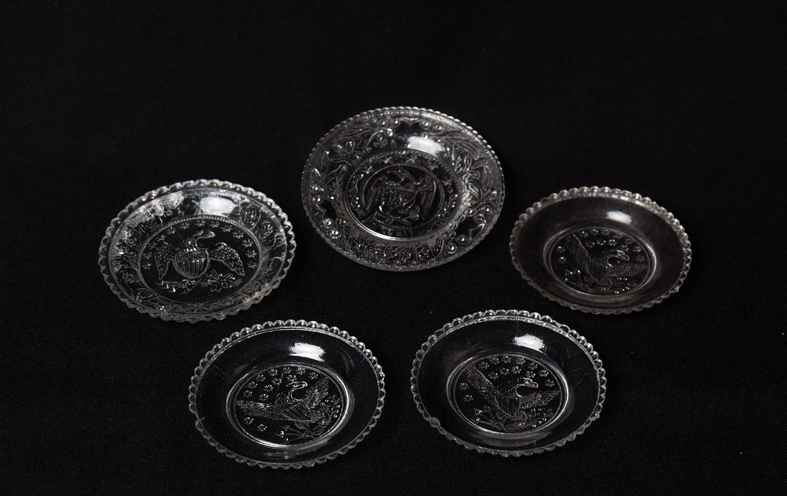 FIVE PRESSED FLINT GLASS EAGLE CUP PLATES.: FIVE PRESSED FLINT GLASS EAGLE CUP PLATES. American, mid- 19th Century. Three are eagles with serrated edges and surrounded with thirteen stars, 3"d., one is Sandwich with central eagle, lacy border,