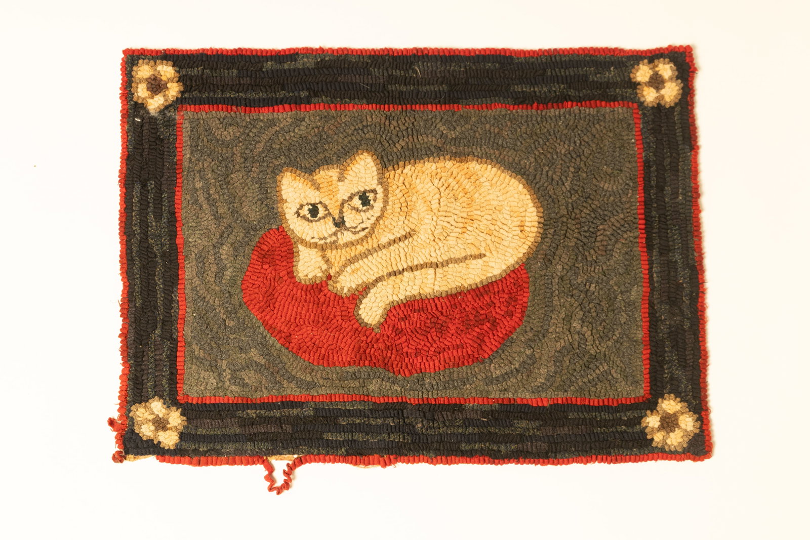 FOLK ART HOOKED RUG WITH RECUMBENT CAT.: FOLK ART HOOKED RUG WITH RECUMBENT CAT. American, Early 20th Century. Rectangular rug depicting a cat curled up on the floor with a gray background, dark border, flowers at corners, and red trim. 22"h