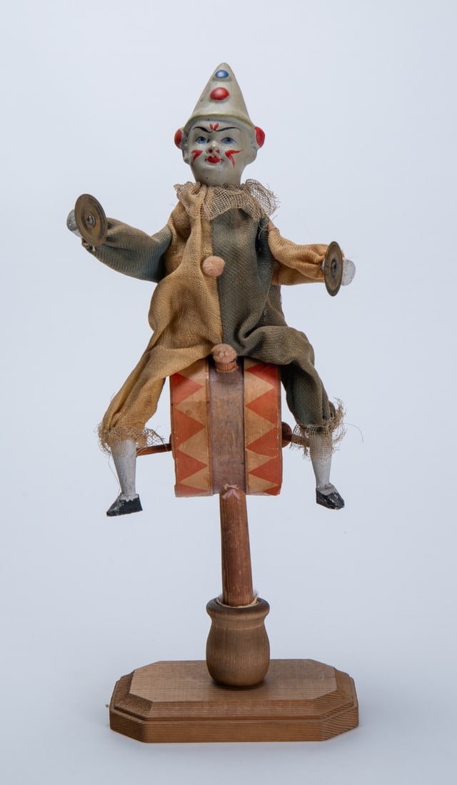 CLOWN ON DRUM CLAPPER TOY.: CLOWN ON DRUM CLAPPER TOY. Germany, late 19th or early 20th Century. Depicting a clown with painted face, cloth clothes, and wood hands and legs sitting atop a drum with brass cymbals; when twisted, i