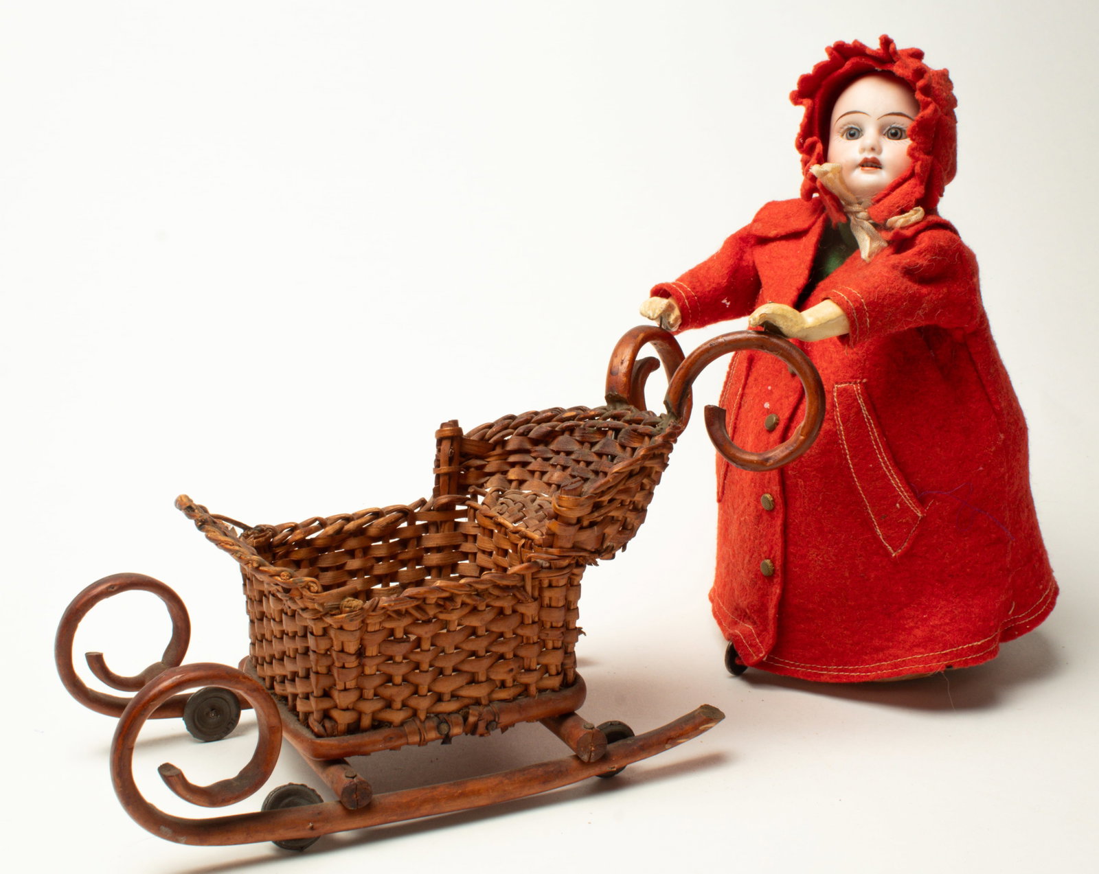 WIND-UP BISQUE HEAD DOLL PUSHING A SLED.: WIND-UP BISQUE HEAD DOLL PUSHING A SLED. Germany, early 20th Century. Girl with a porcelain face, glass eyes, and open mouth wearing a red hooded coat, with four brass buttons. Her hood is tied under