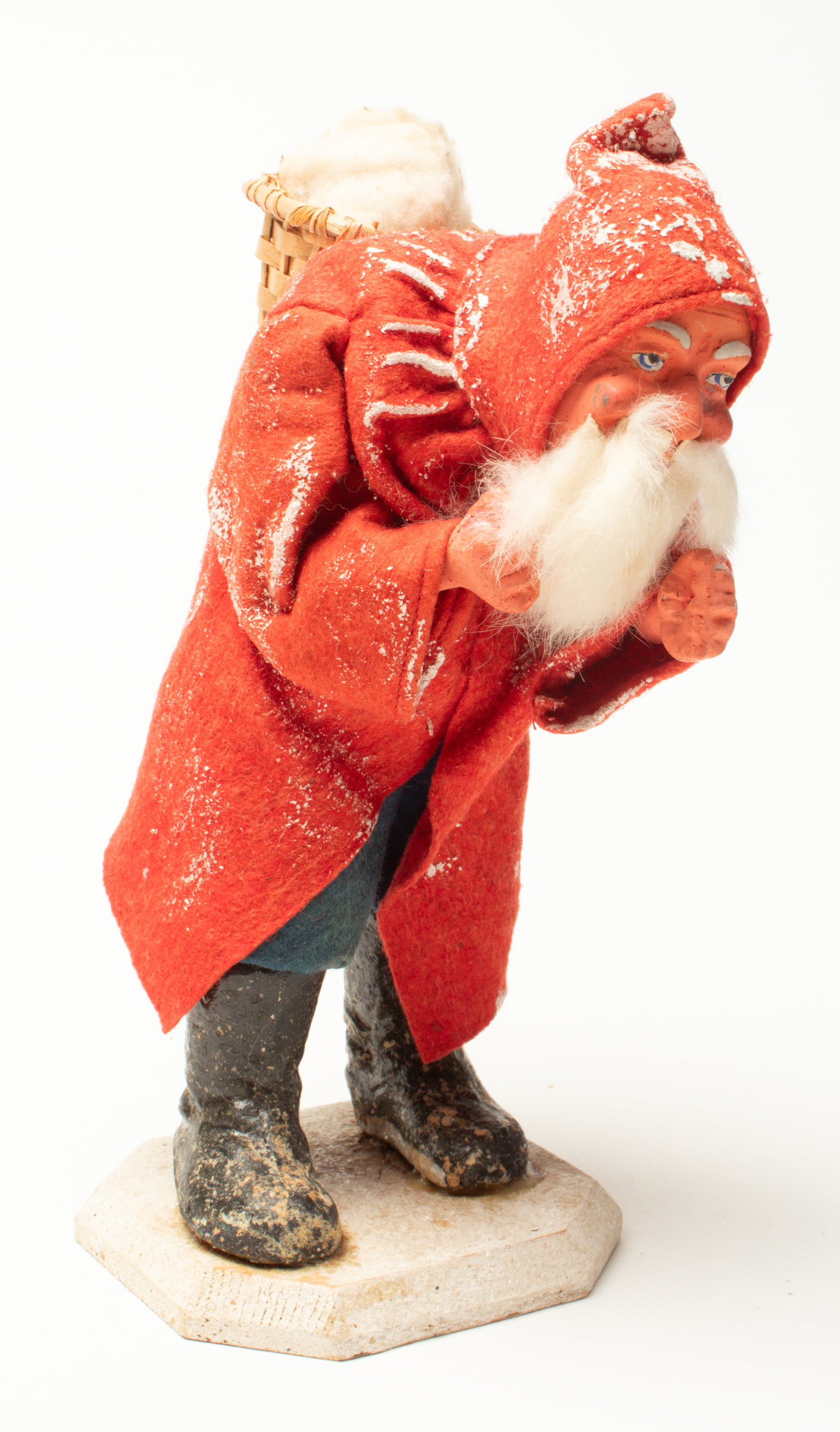 GERMAN TRUDGING SANTA FIGURE.: GERMAN TRUDGING SANTA FIGURE. Germany, early 20th Century. A slumped over Santa wearing a hooded red coat with dustings of snow, blue paints, and black boots with a basket on his back. On a wooden bas