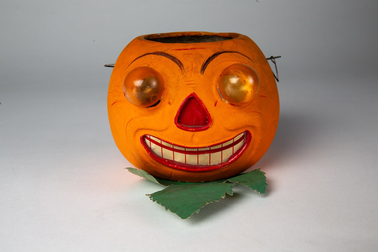 HALLOWEEN JACK-O-LANTERN WITH BLOWN GLASS EYES.: HALLOWEEN JACK-O-LANTERN WITH BLOWN GLASS EYES. Germany, early 20th Century. The composition lantern with blown, amber glass eyes and smiling mouth; on a paper leaf base; 4.25"h. 5"w. Provenance: