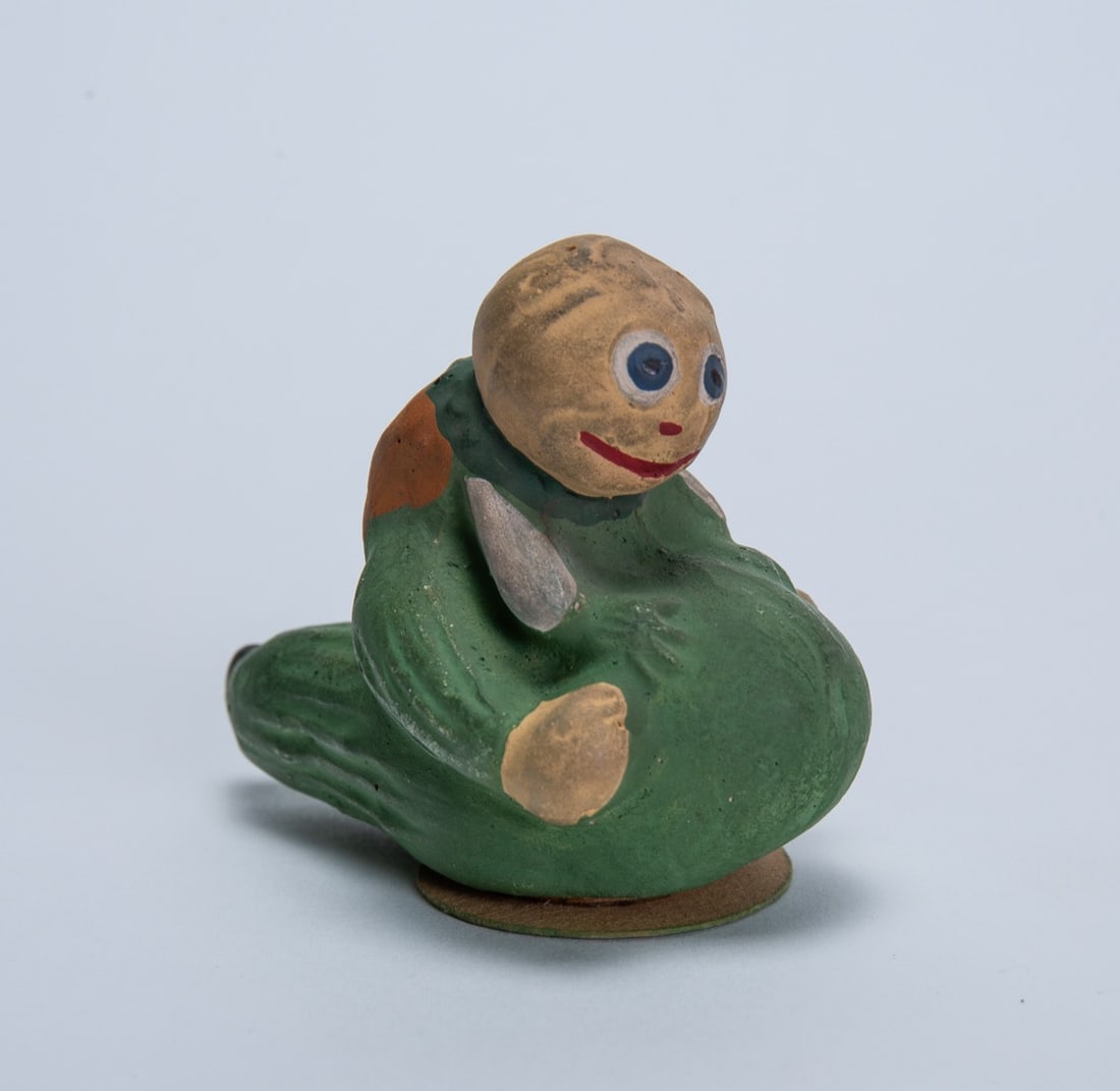 PUMPKIN-HEADED VEGGIE MAN HALLOWEEN CANDY CONTAINER.: PUMPKIN-HEADED VEGGIE MAN HALLOWEEN CANDY CONTAINER. Germany, early 20th Century. Papier-mache. Depicting a pumpkin-headed figure riding a green squash; 2.25"h. 2.75"w. Provenance: Nancy Goff