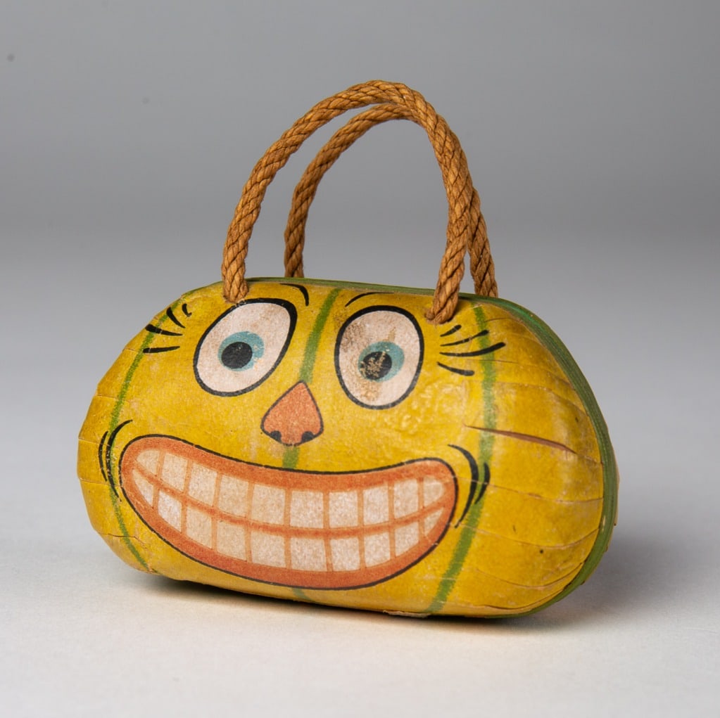 HALLOWEEN PURSE-SHAPED CANDY CONTAINER WITH HANDLE.: HALLOWEEN PURSE-SHAPED CANDY CONTAINER WITH HANDLE. Germany, 20th Century. Having a jack-o-lantern face on one side with melon stripes on both; interior with fleur-de-lis paper; 3"h. 2.5"w. .