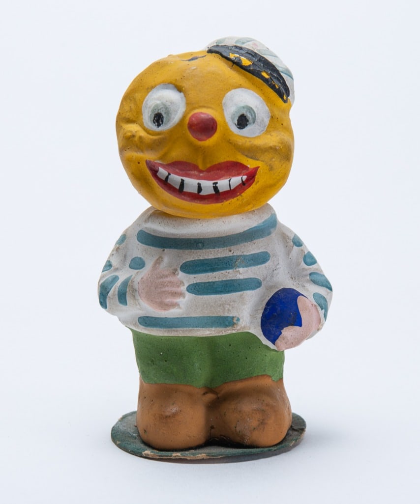 PUMPKIN BOY BASEBALL PLAYER HALLOWEEN CANDY CONTAINER.: PUMPKIN BOY BASEBALL PLAYER HALLOWEEN CANDY CONTAINER. Germany, 20th Century. Composition. The smiling pumpkin-headed boy wearing a striped cap and shirt with green pants while holding a ball; 3.5"h.