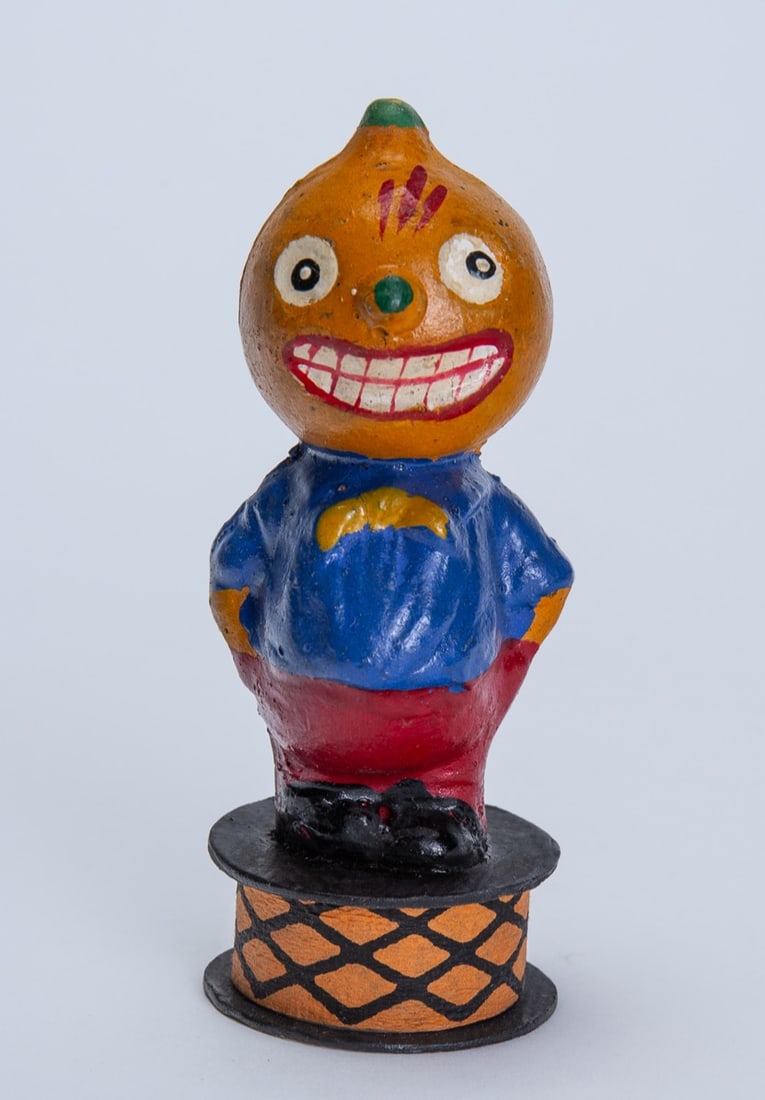PUMPKIN MAN HALLOWEEN CANDY CONTAINER.: PUMPKIN MAN HALLOWEEN CANDY CONTAINER. Germany, 20th Century. Composition. Smiling pumpkin-headed man wearing a blue shirt and red pants with hands in pockets; on a crepe paper base; 4.5"h. 2"w.