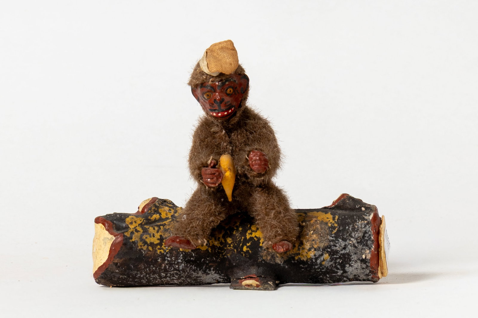 MONKEY ON LOG CANDY CONTAINER.: MONKEY ON LOG CANDY CONTAINER. Germany, early 20th Century. A monkey with mohair fur, glass eyes, painted face, wearing a cap, sitting on a painted, papier-mache log holding a banana in its hand.