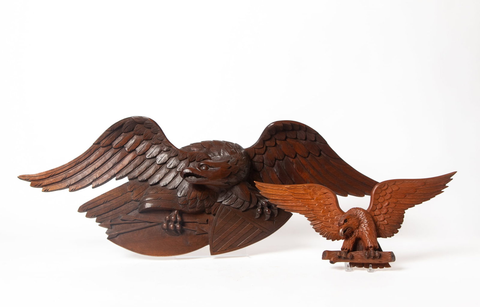 TWO CARVED SPREAD-WINGED EAGLES.: TWO CARVED SPREAD-WINGED EAGLES. American, 19th and 20th Century. The largest is walnut, spread-winged, clutching a shield and arrows in his talons, 15"h. 44"w. And the second is pine, spread-winged
