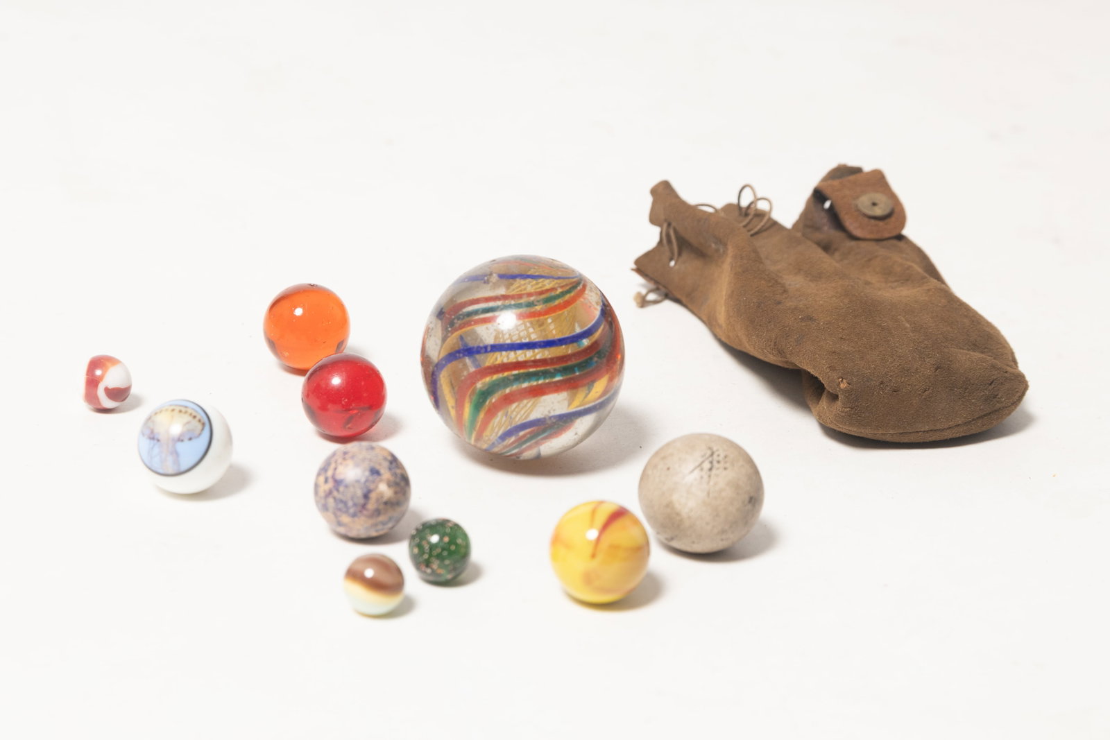 COLLECTION OF TEN MARBLES, INCLUDING LARGE LATTICINO CORE SWIRL GLASS MARBLE AND LEATHER MARBLE BAG.: COLLECTION OF TEN MARBLES, INCLUDING LARGE LATTICINO CORE SWIRL GLASS MARBLE AND LEATHER MARBLE BAG. German and American, late 19th Century. Large handmade marble with yellow core and blue, green, red